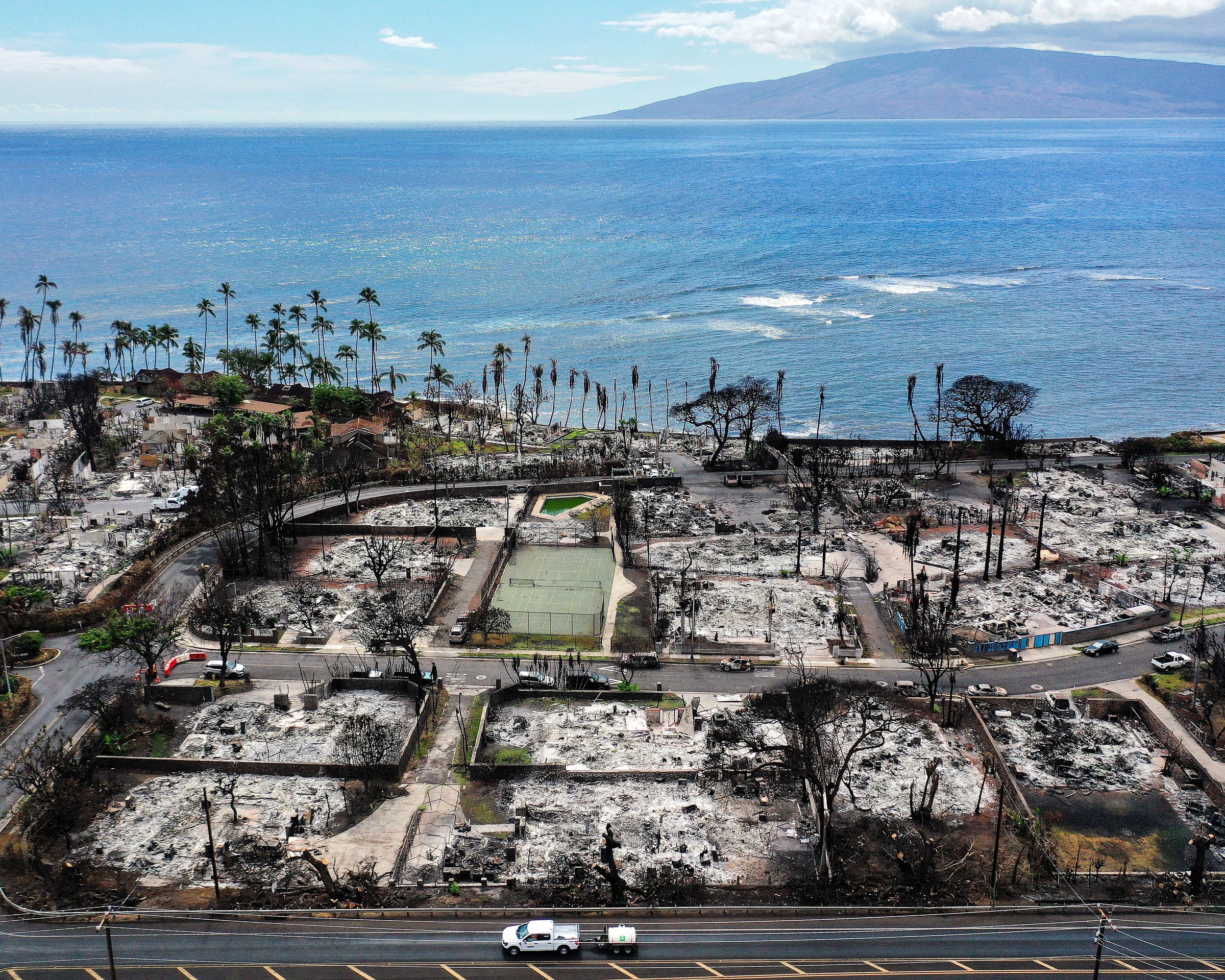 In an aerial view, a recovery vehicle drives past burned structures and cars in a neighborhood next to shoreline.