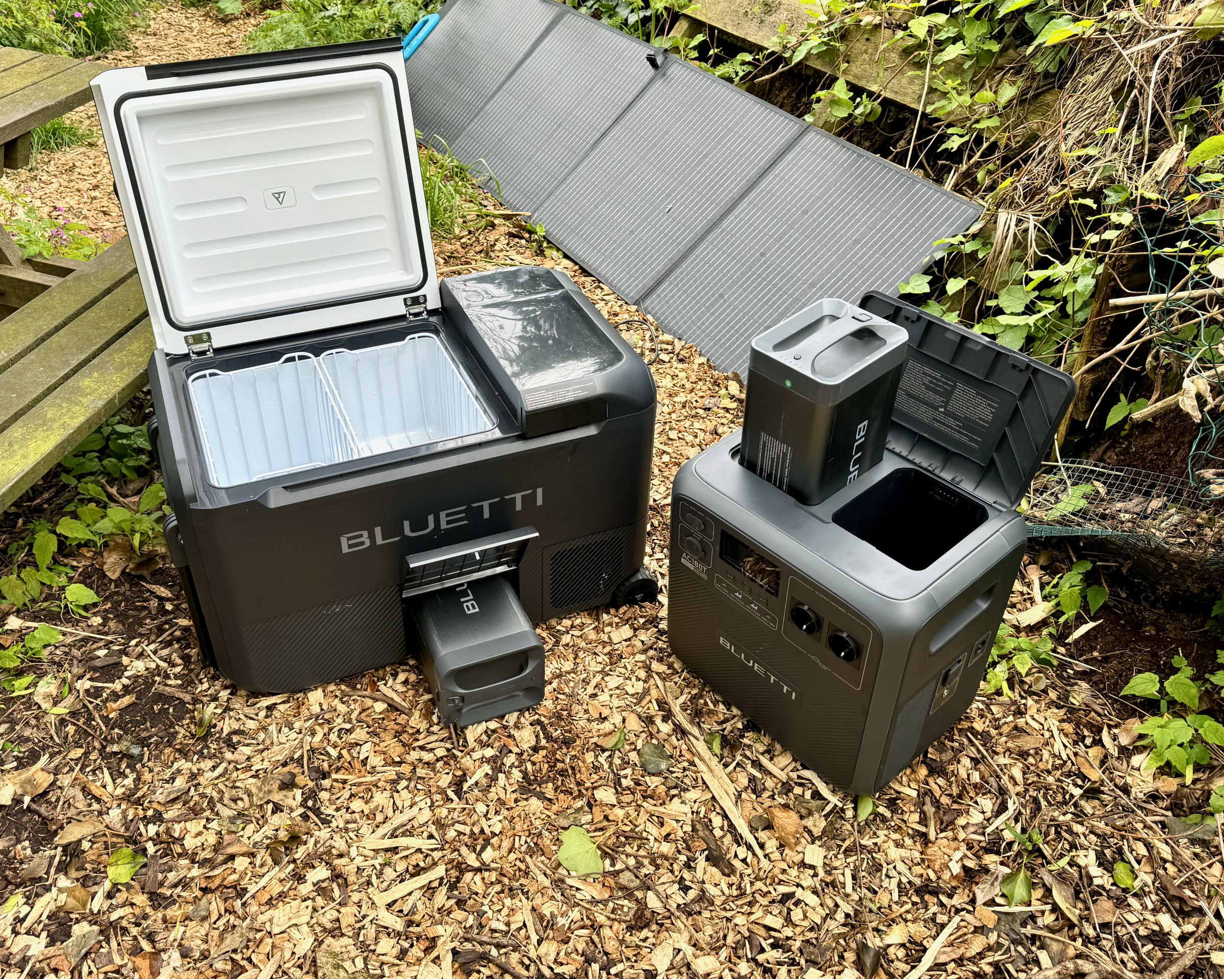 The Bluetti MultiCooler and AC180T solar generator sitting in a park next to a picnic table with a solar panel in the background.
