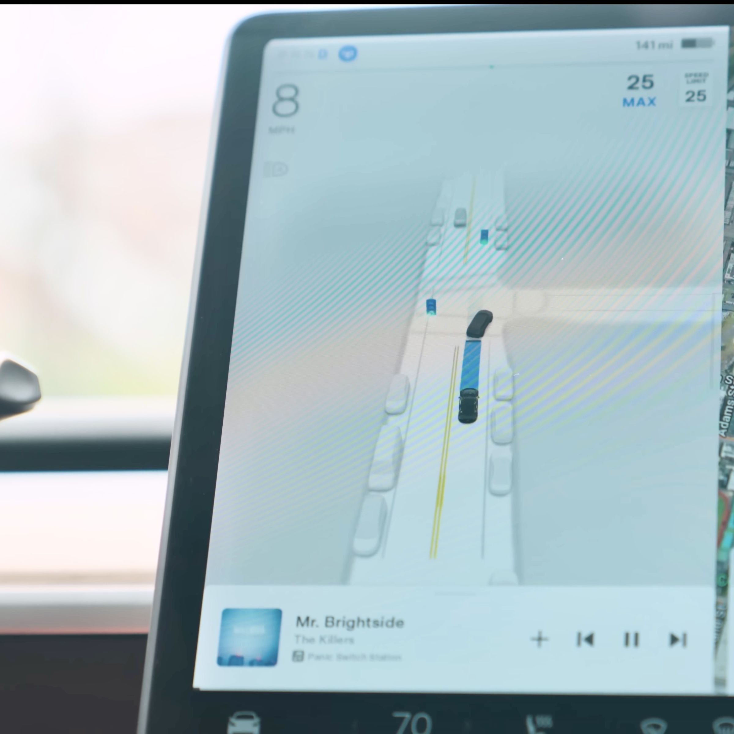 Left side of Tesla Model 3 main screen showing a computer-generated image of an intersection with cars parked on the sides and the Model 3 following another car