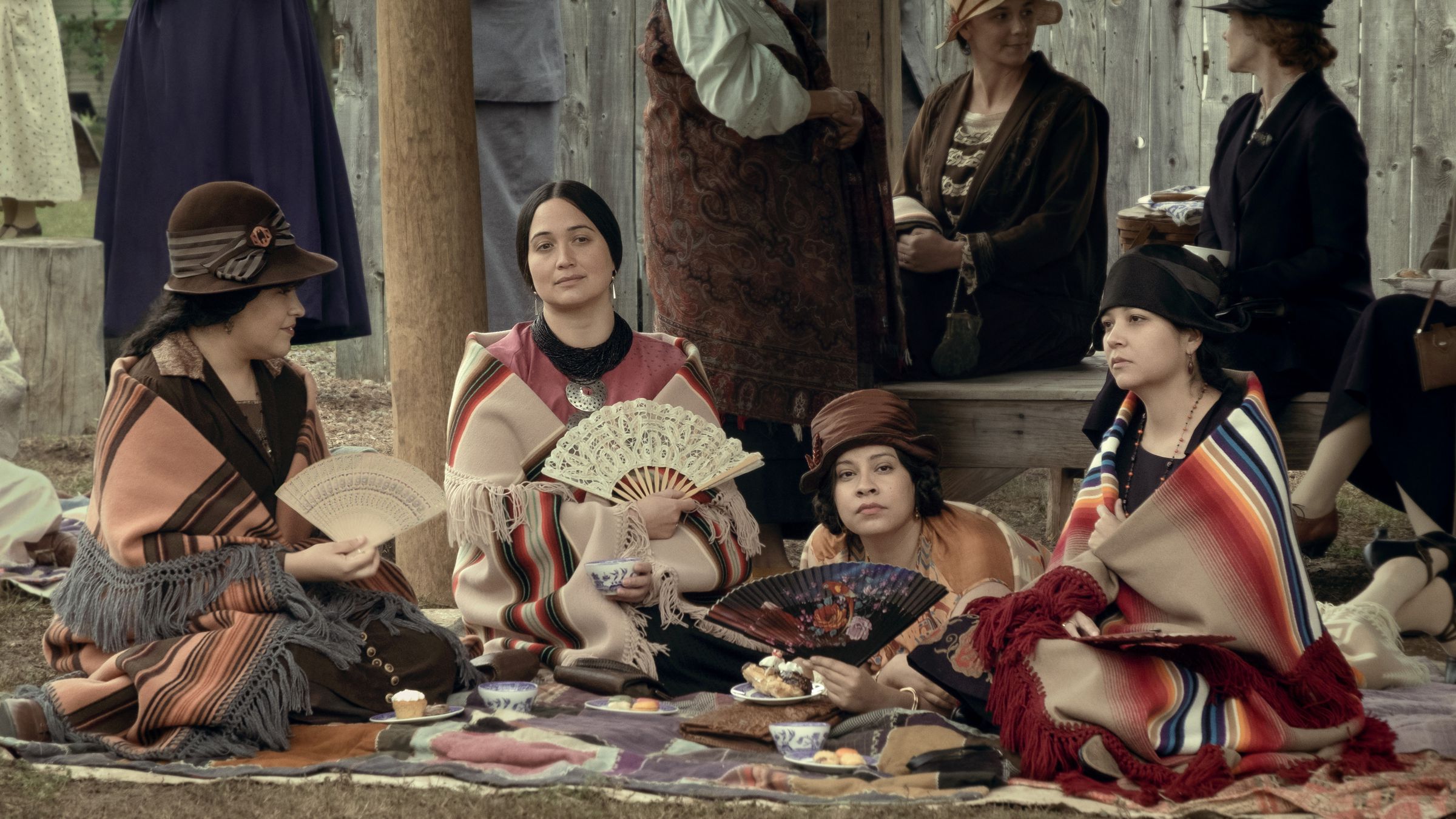 A group of four Native American women sitting together on the ground in front of a store while they fan themselves and enjoy pastries.