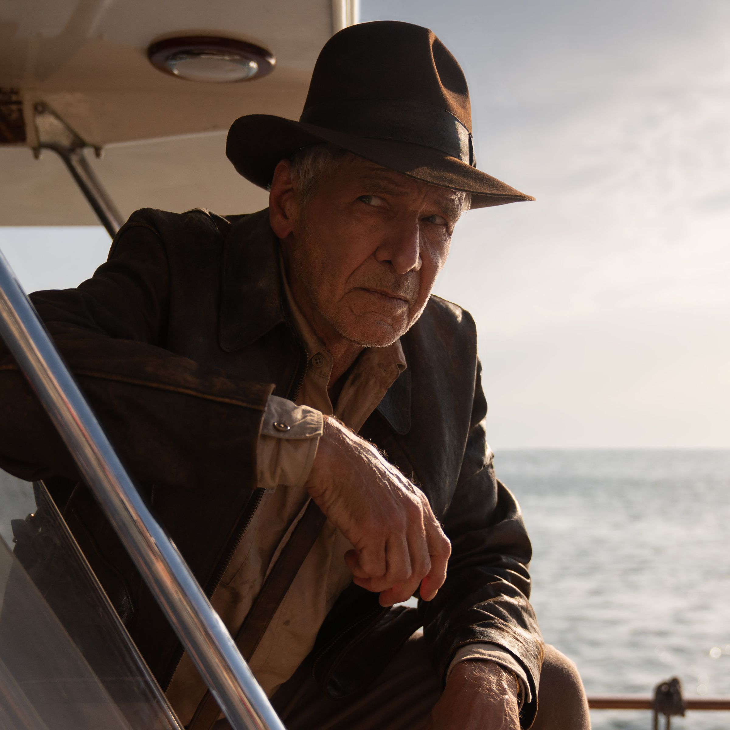 A mean wearing a fedora, shirt, and jacket. The man is leaning out of an opening on the deck of a boat.