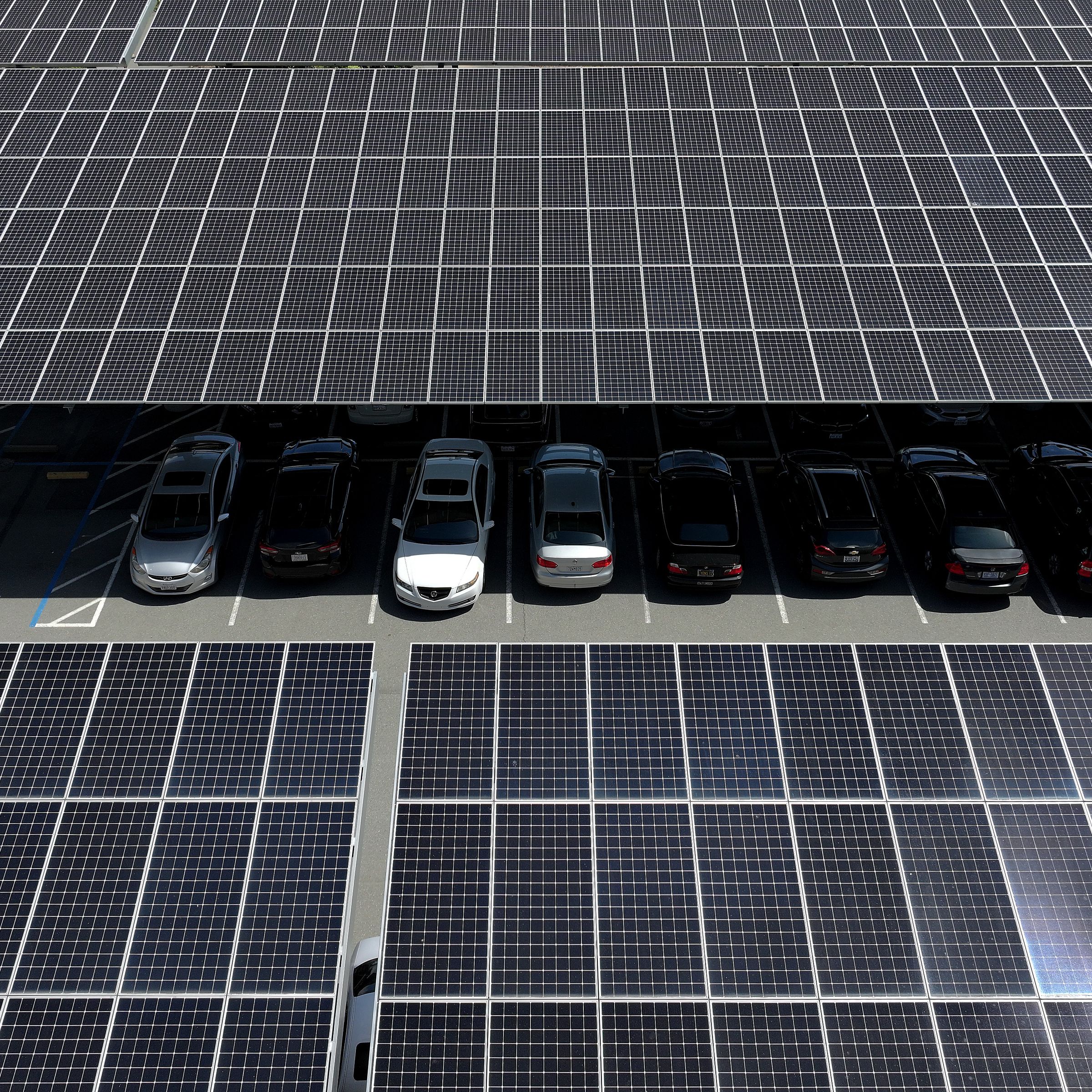 In an aerial view, solar panels are seen in a parking lot shading cars parked underneath them.