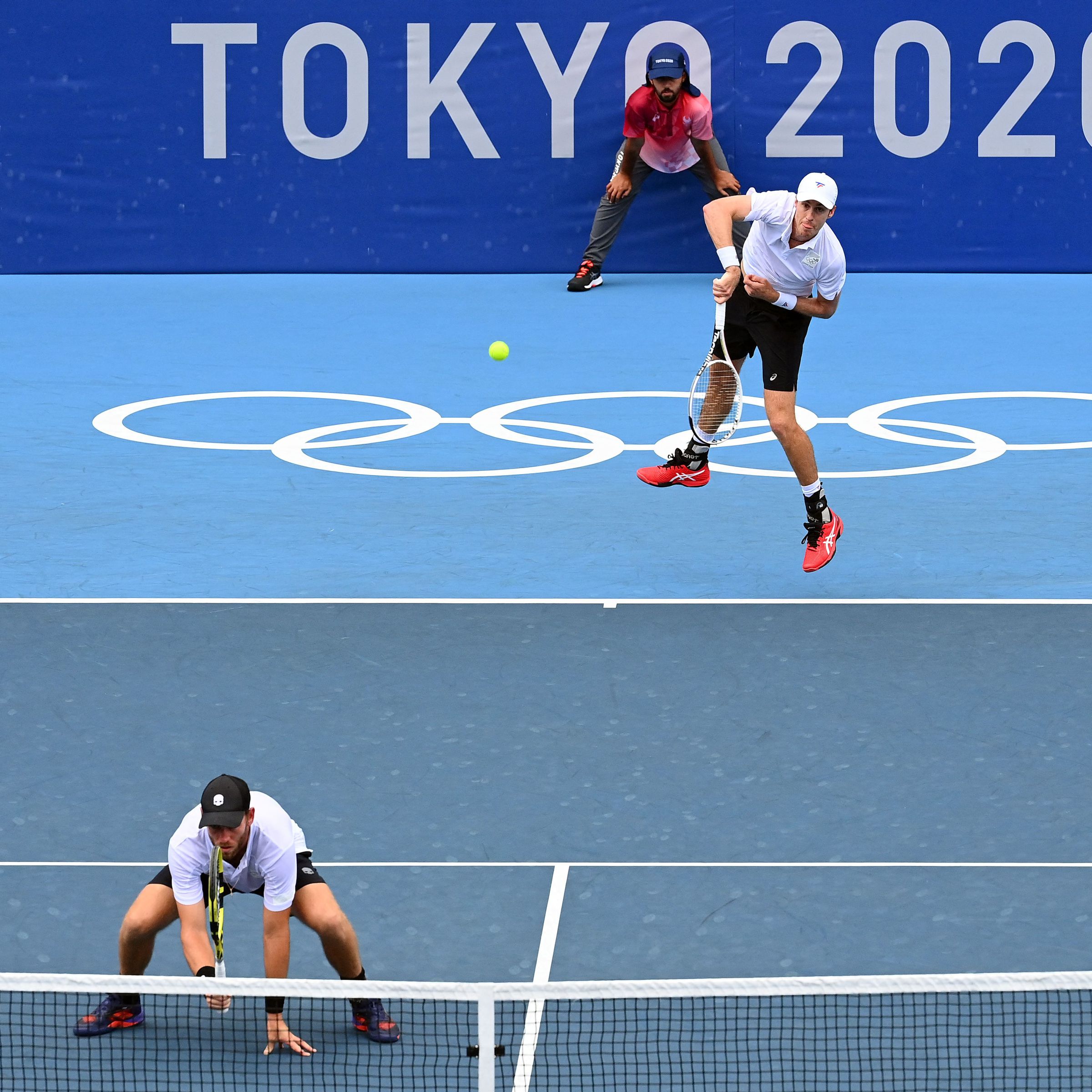 A man leaps to hit a tennis ball on an Olympic court. Another man is crouching on the court in front of him.