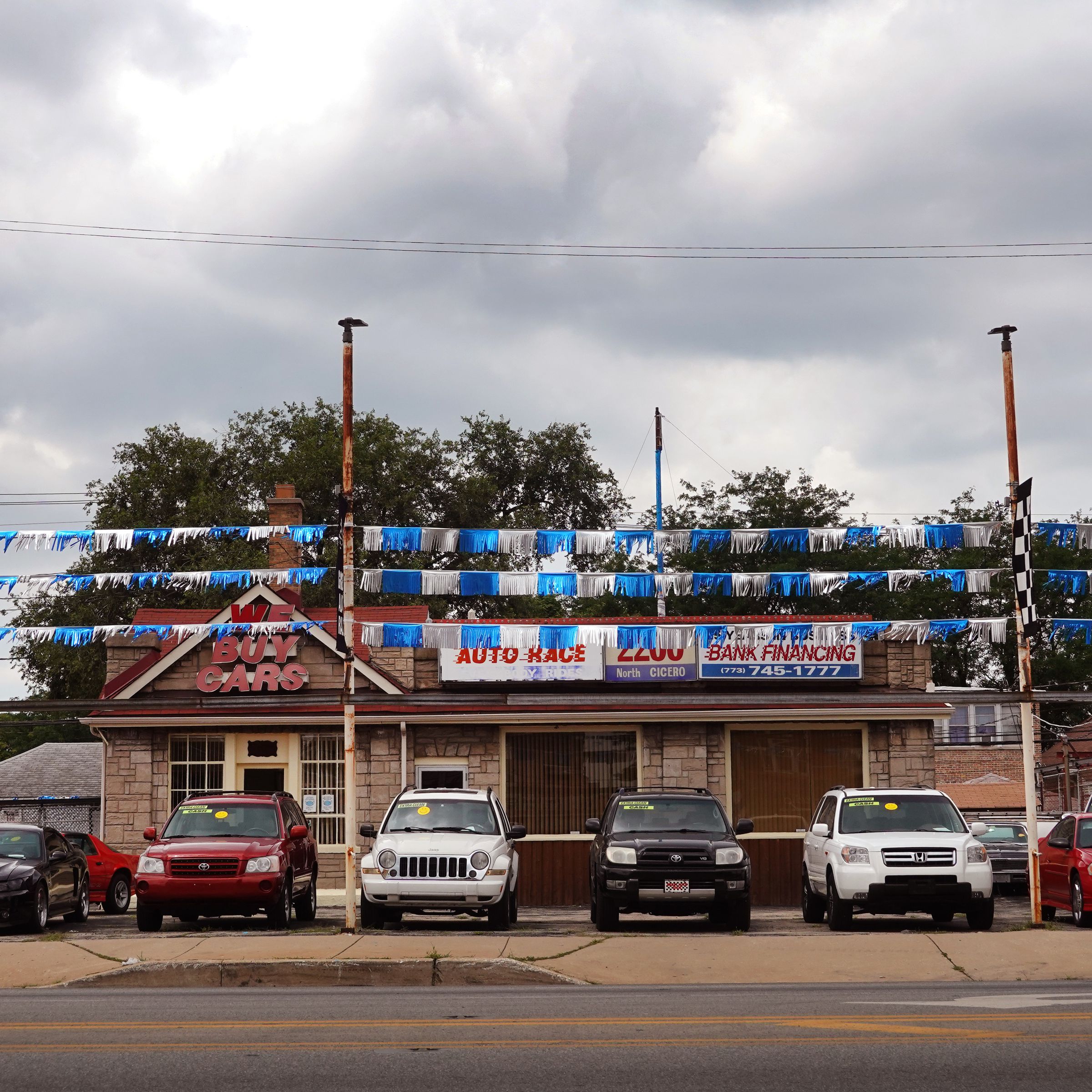 A picture of a car dealership showing the front of several cars in a lot, three blue and silver lines of streamers strung from poles above, and a small building behind them, on a cloudy day.