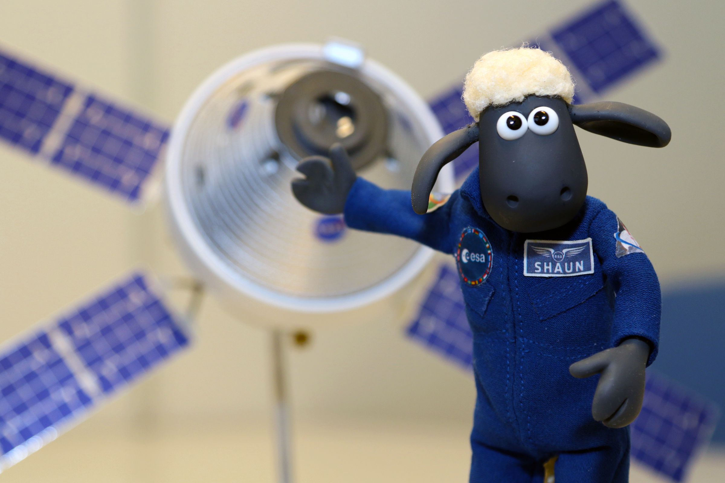 A plush cartoon sheep holds out a hoof towards a small model of the Orion capsule.
