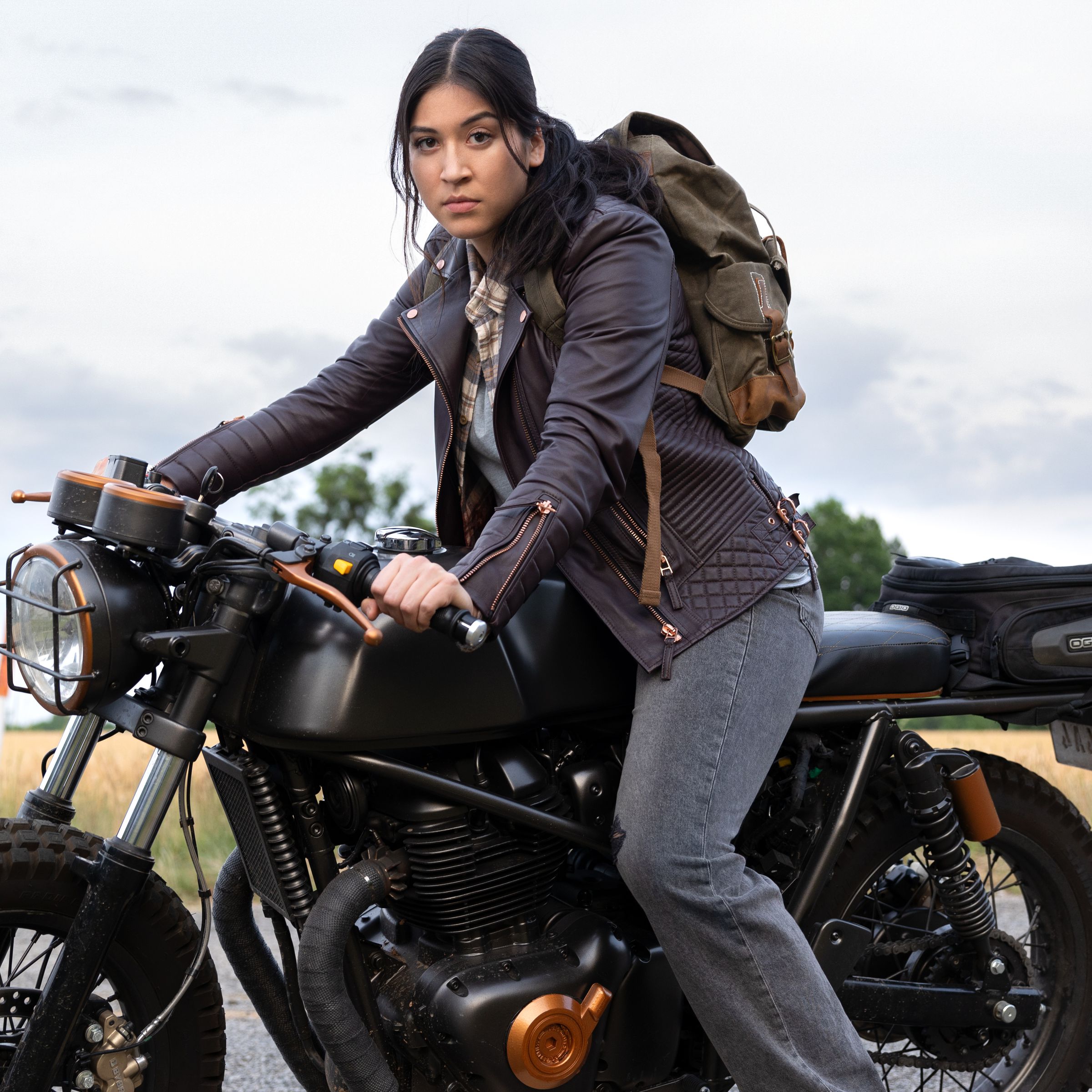A wide shot of a woman with long black hair tied back in a pony tail, wearing a leather jacket, jeans, and straddling a motor cycle.