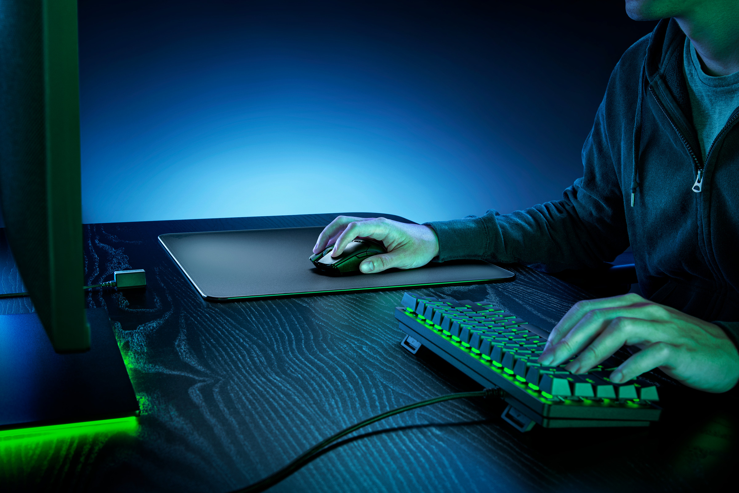A user clicks on a mouse atop the Razer Atlas with their right hand and uses a green-lit mechanical keyboard with their left.