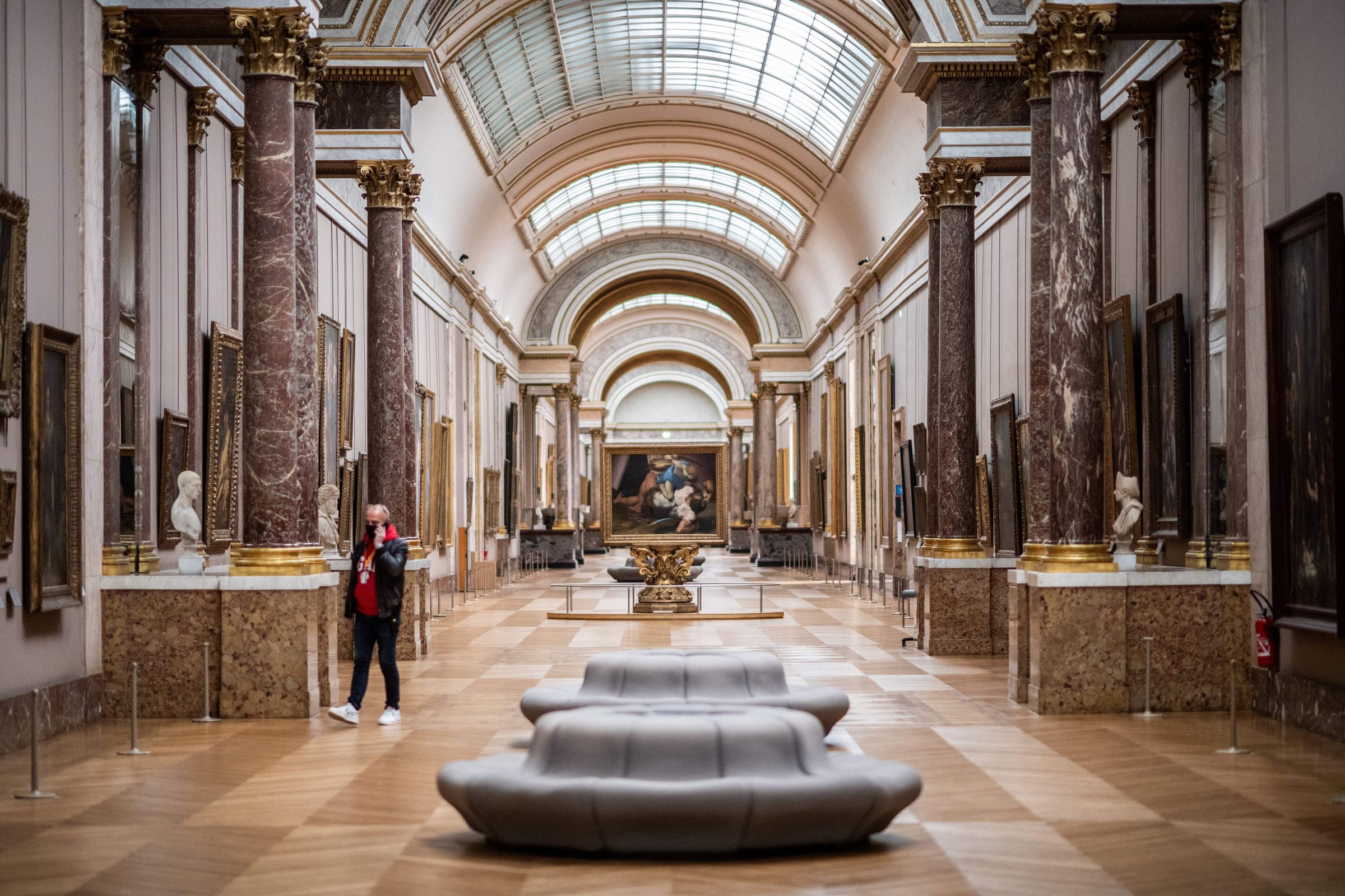 A spacious, sunlit gallery hall at the Louvre, with skylights, marble pillars, and artworks along the walls. 