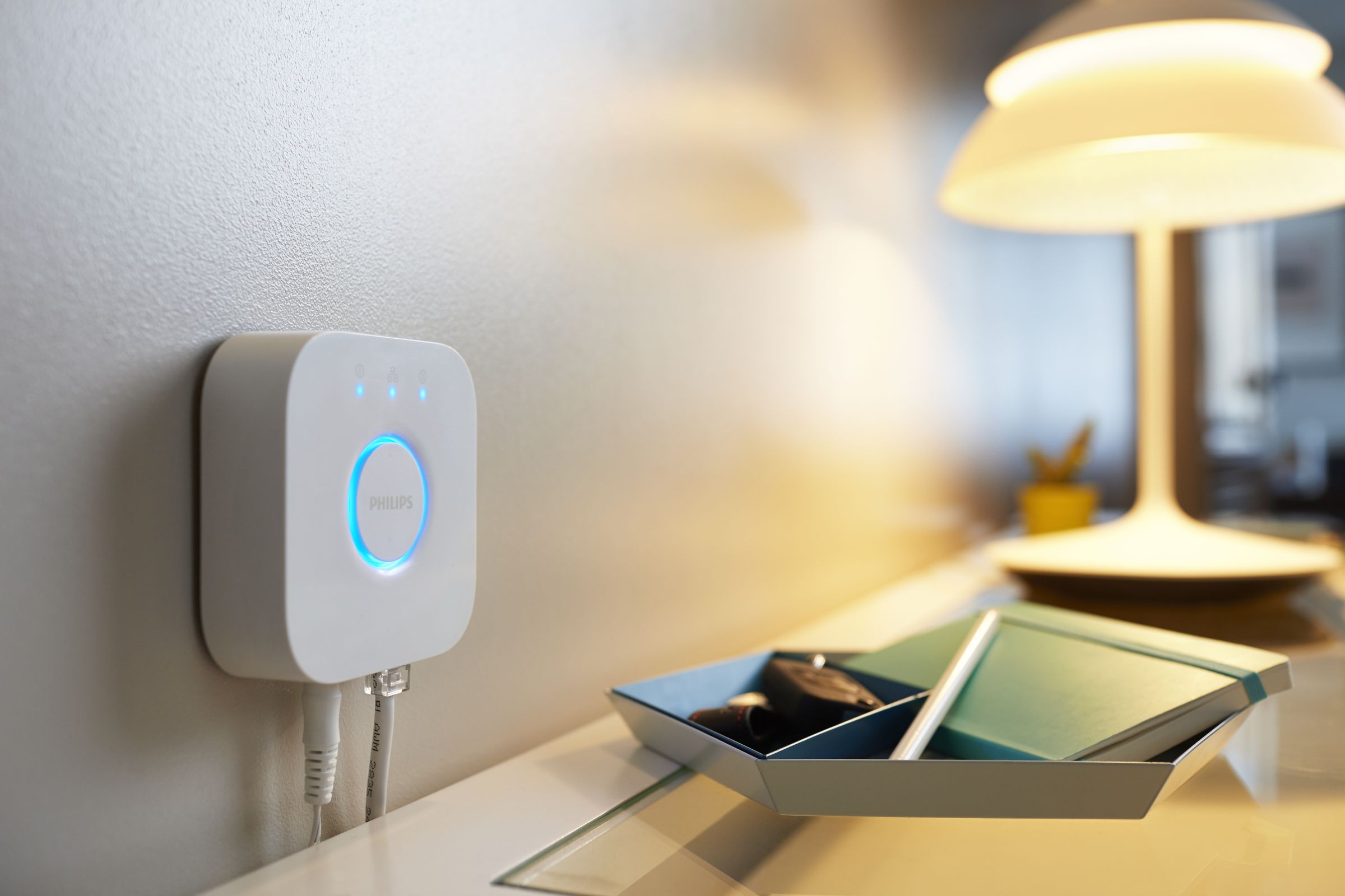 Philips Hue Bridge device shown wall-mounted above a table near a lamp.