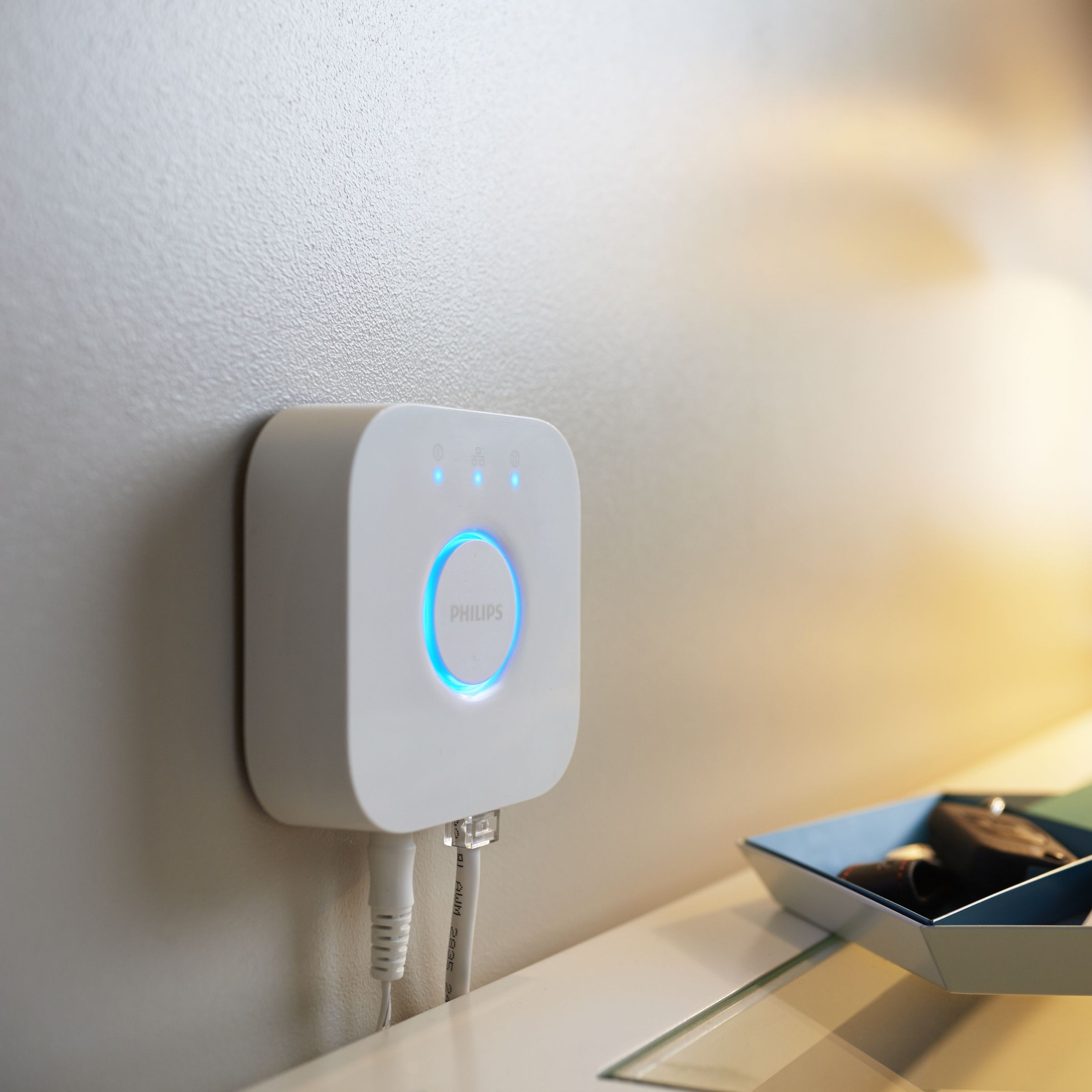 Philips Hue Bridge device shown wall-mounted above a table, near a lamp.