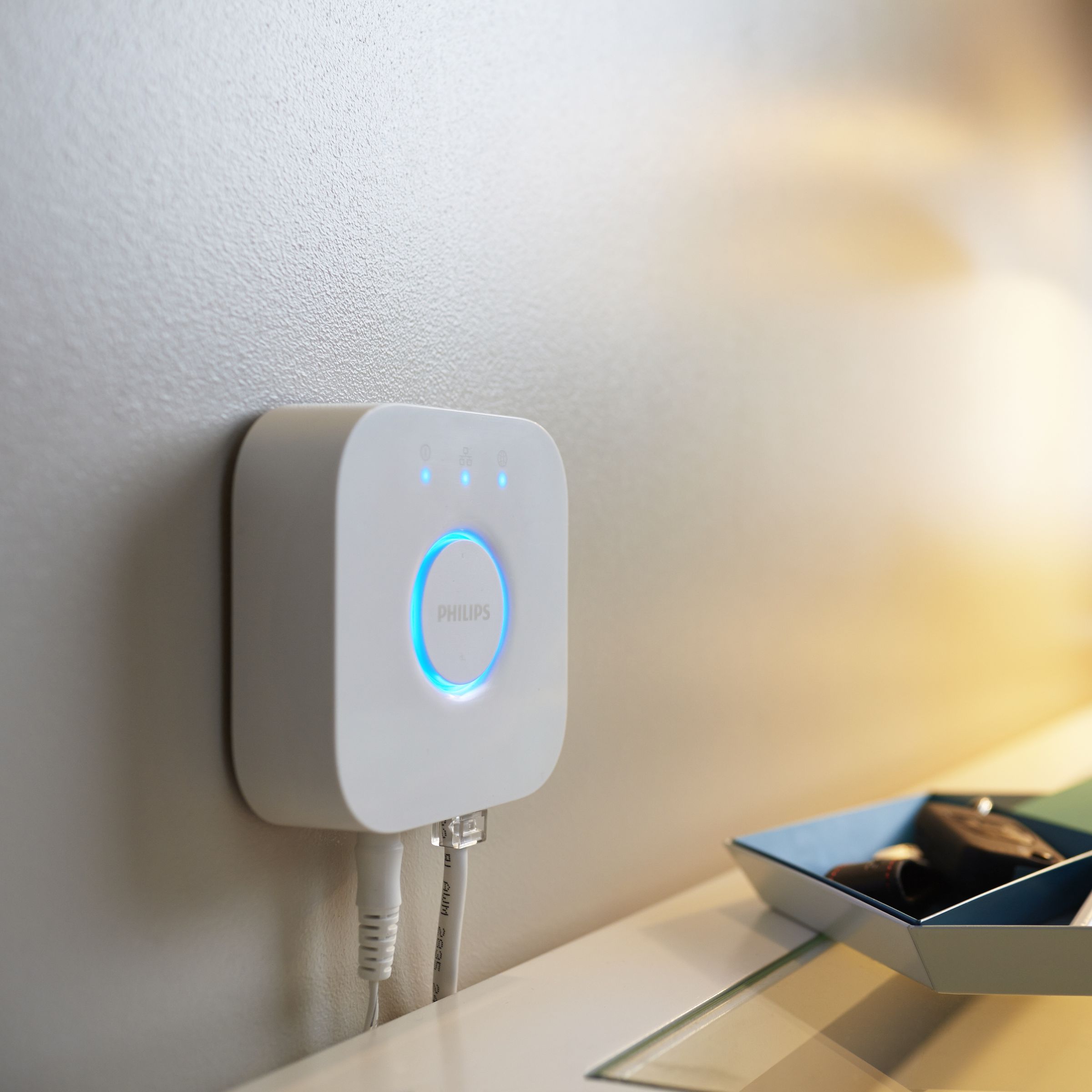 Philips Hue Bridge device shown wall-mounted above a table near a lamp.