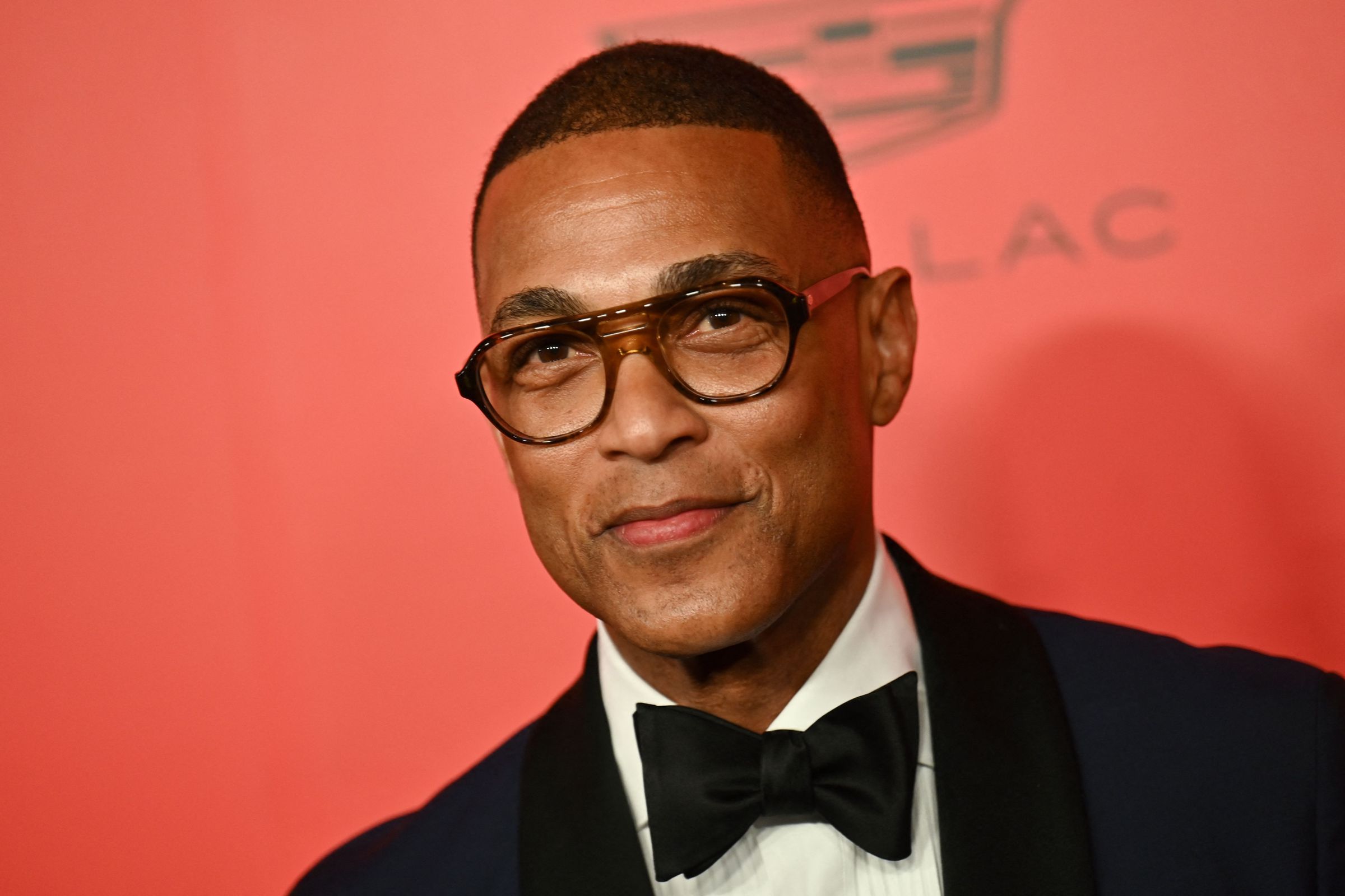 A tight shot of a man in a tuxedo wearing glasses and a bowtie. Behind the man is a red backsplash.