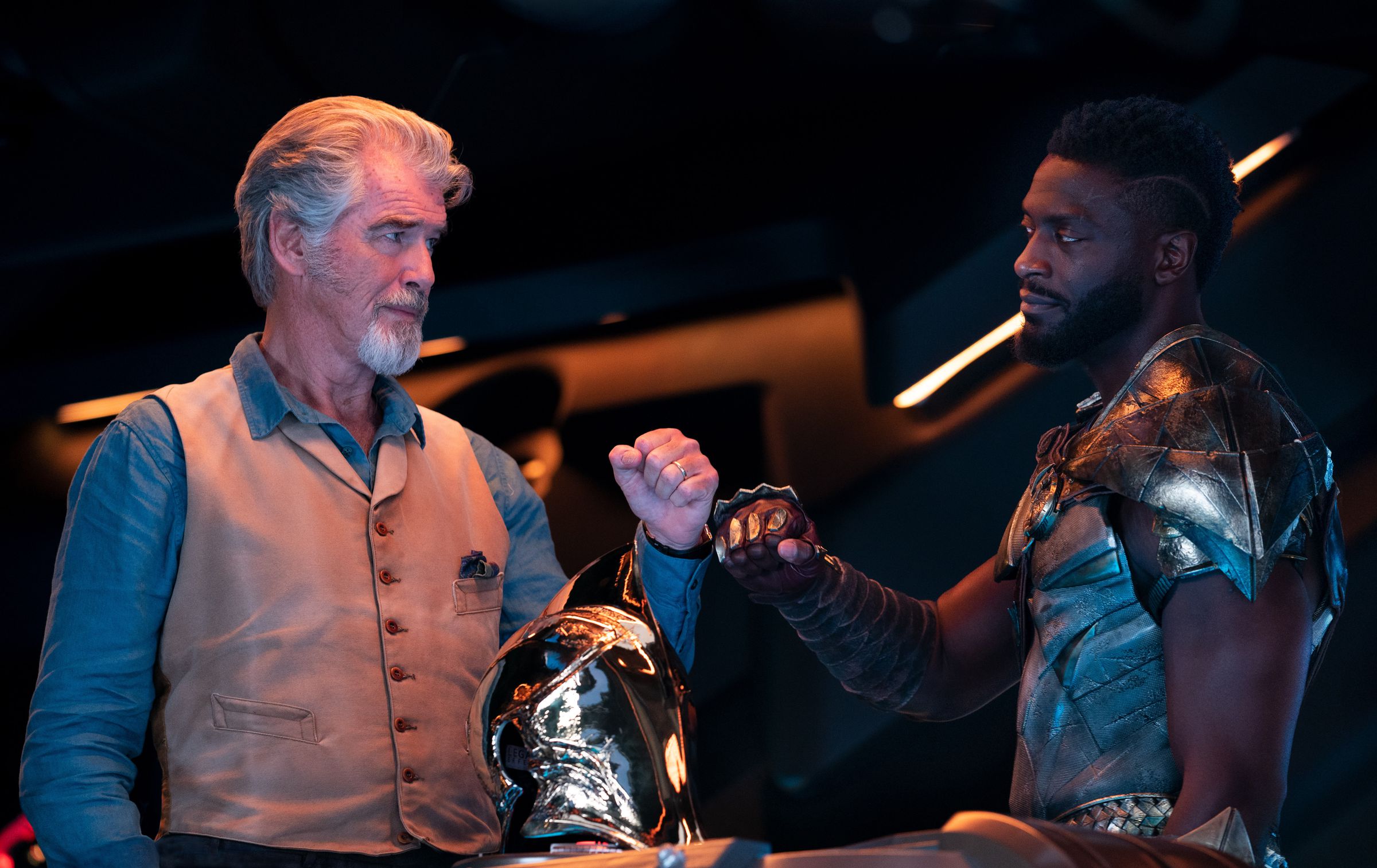 A graying old man wearing a blue shirt and a tan vest bumps fists with a younger man wearing golden armor that’s vaguely bird-themed. In the foreground sits a shining golden helmet reflecting light from throughout the room.