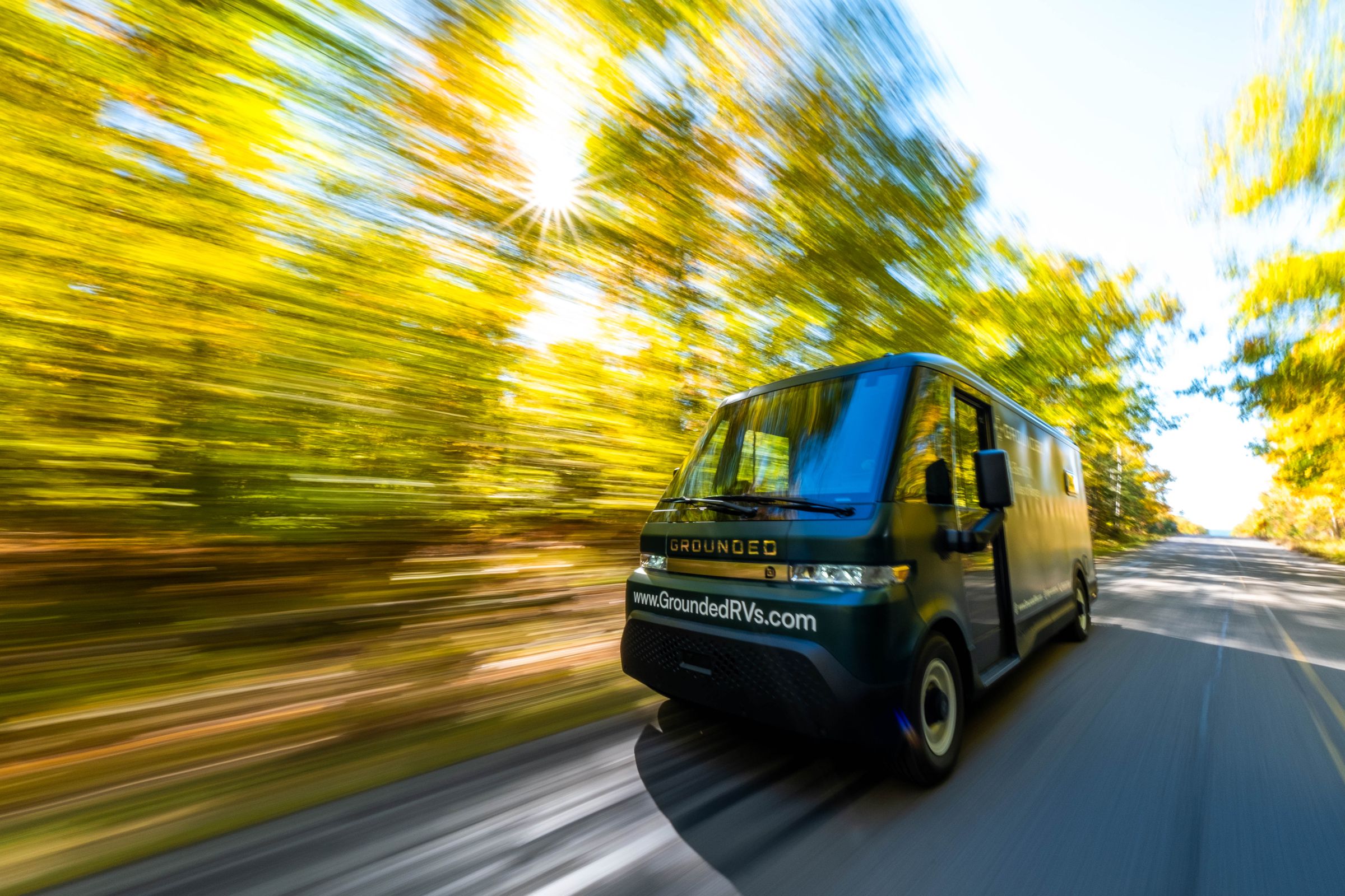 A boxy Grounded G2 camper van drives down a blurry tree-lined road.