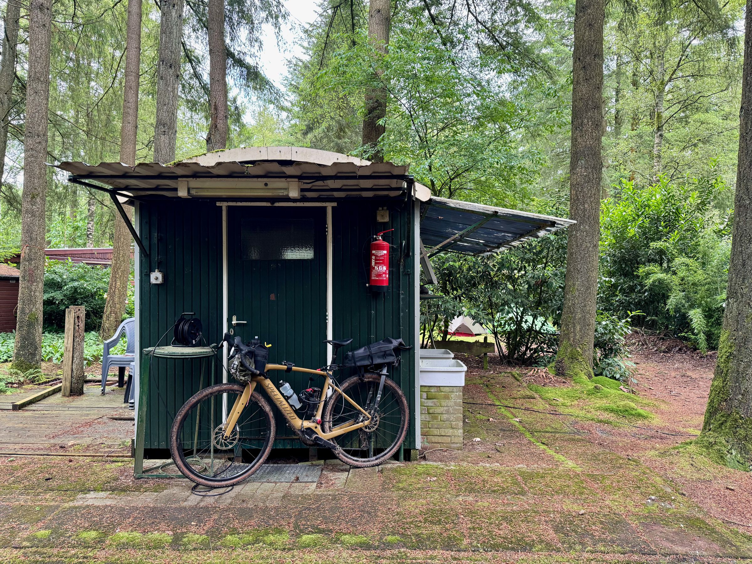 <em>This campsite ran an extension cord out from the shed to charge the e-bike. The overhang gave it some protection from the elements. You can see our tents directly behind the shed.</em>