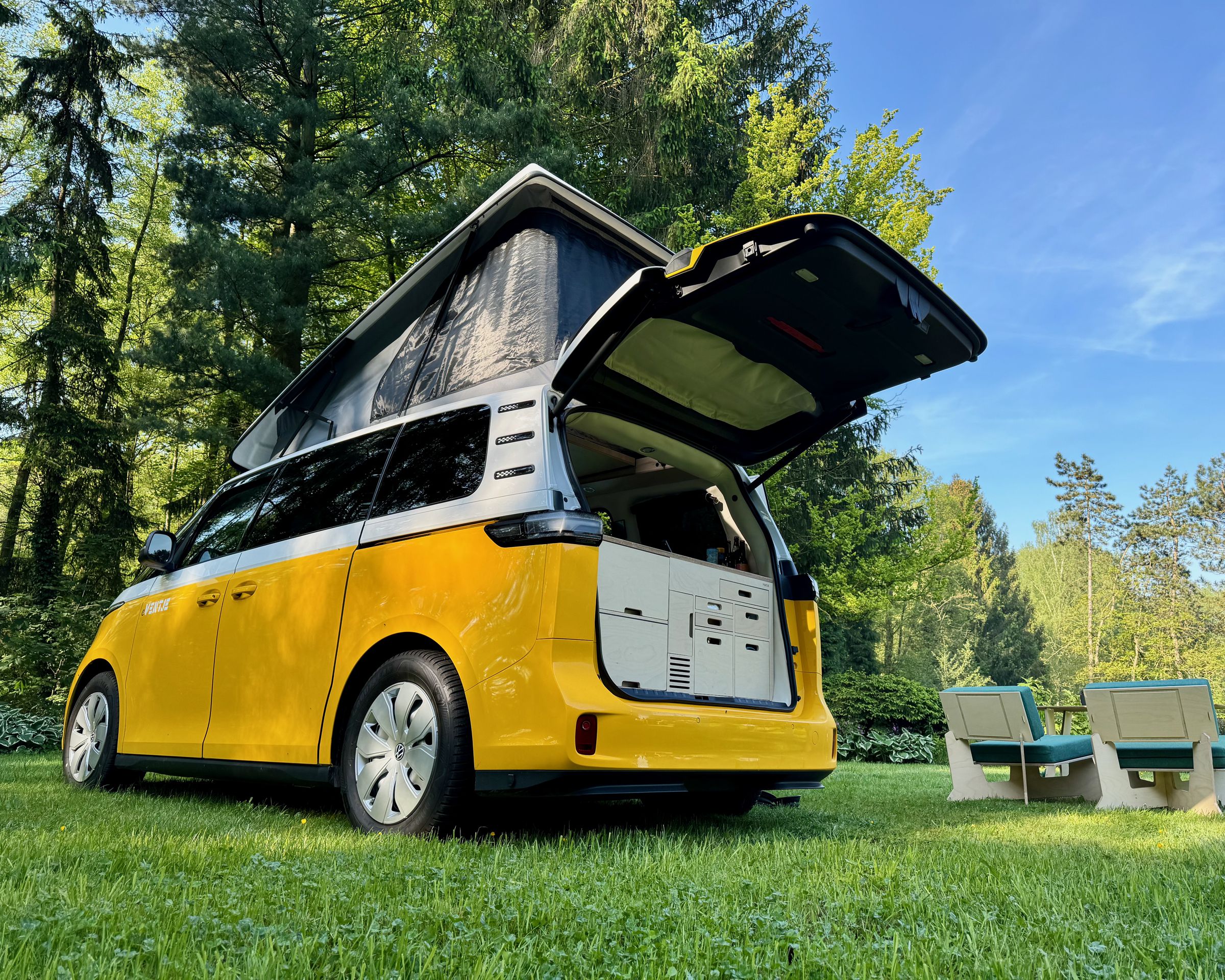A customized ID Buzz Ventje sits on the grass beneath the trees with the pop-top and tail gate open and furniture setup on the lawn.