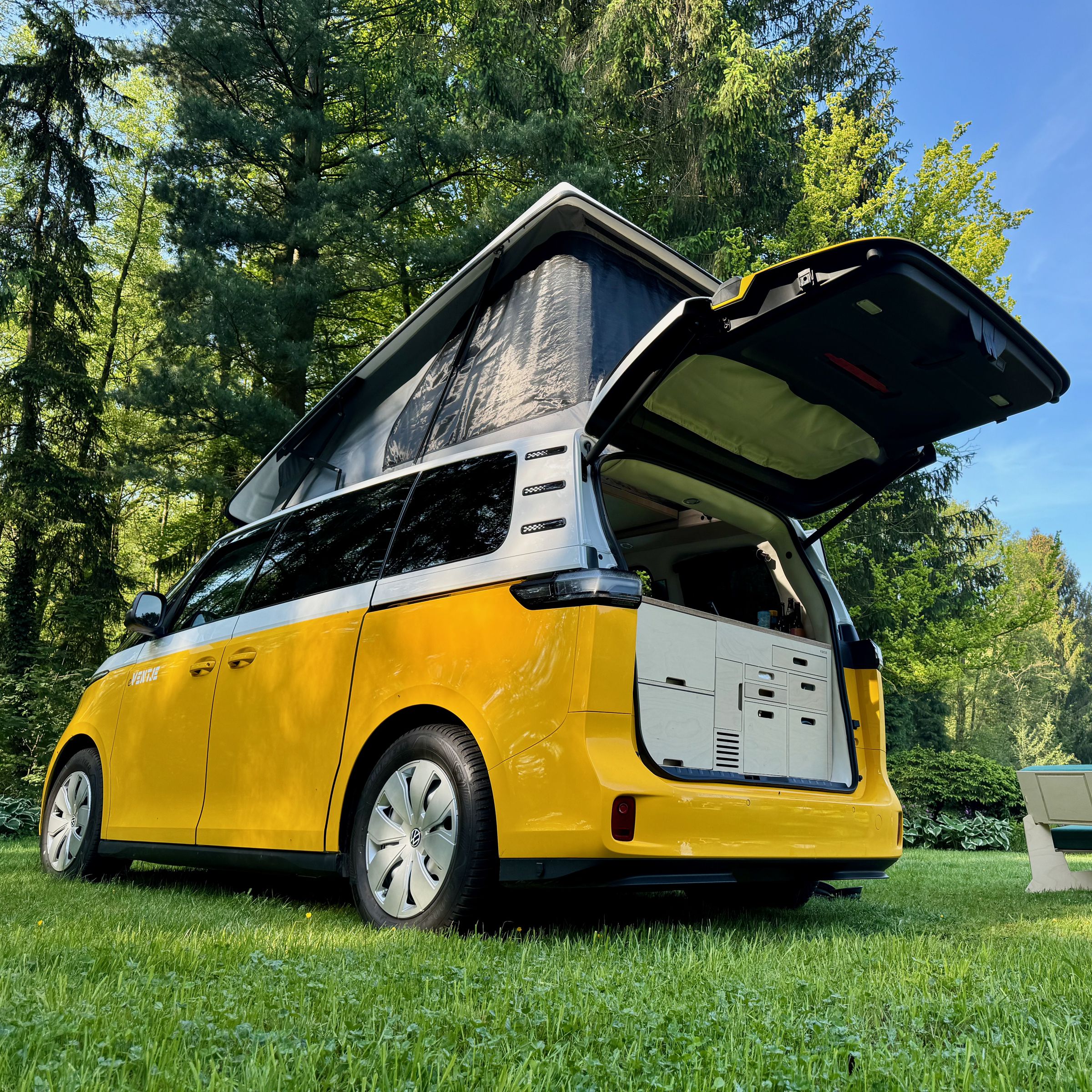 A customized ID Buzz Ventje sits on the grass beneath the trees with the pop-top and tail gate open and furniture setup on the lawn.