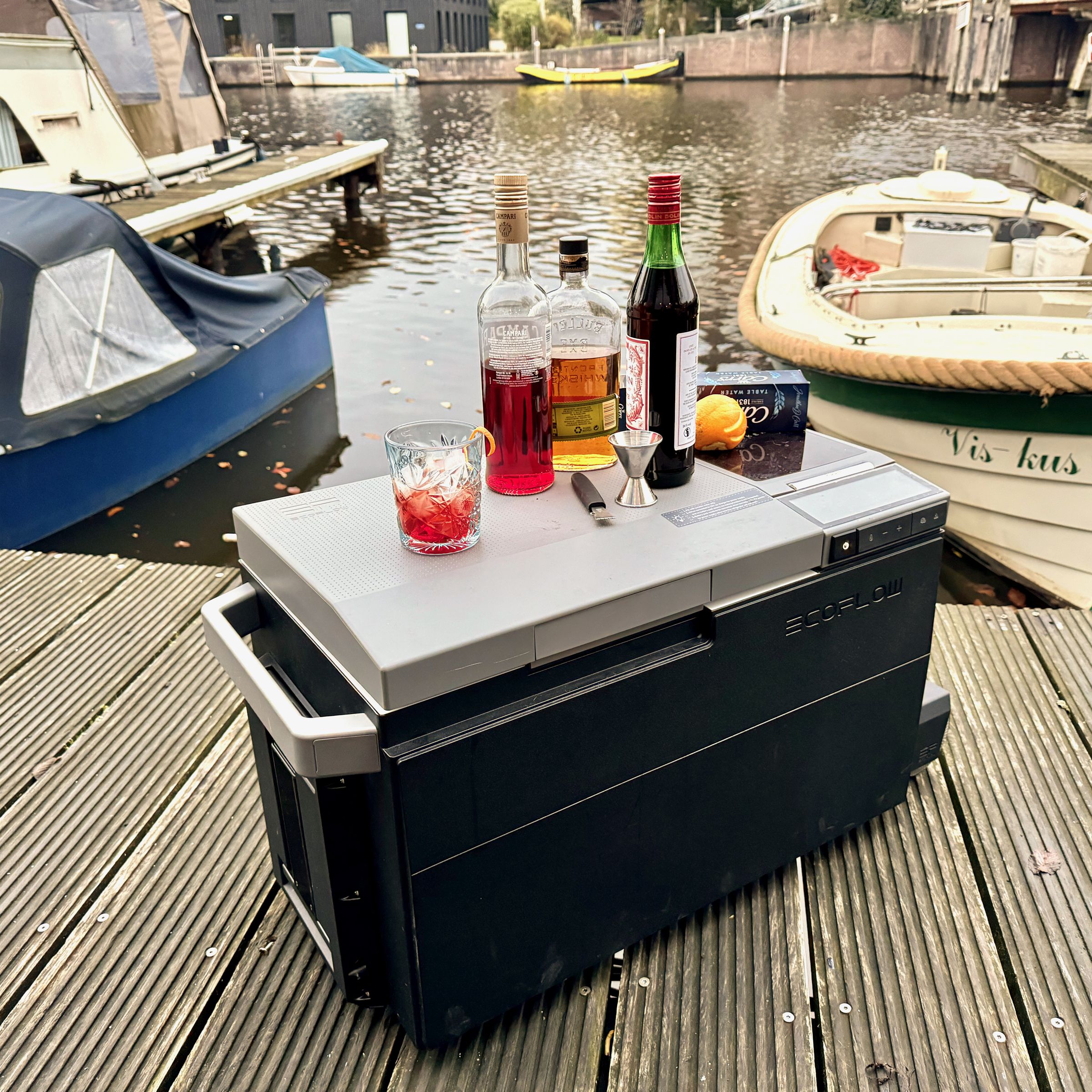 EcoFlow Glacier sits on a dock with boats in the background. On top of the closed lid are ingredients to make a boulevardier cocktail.