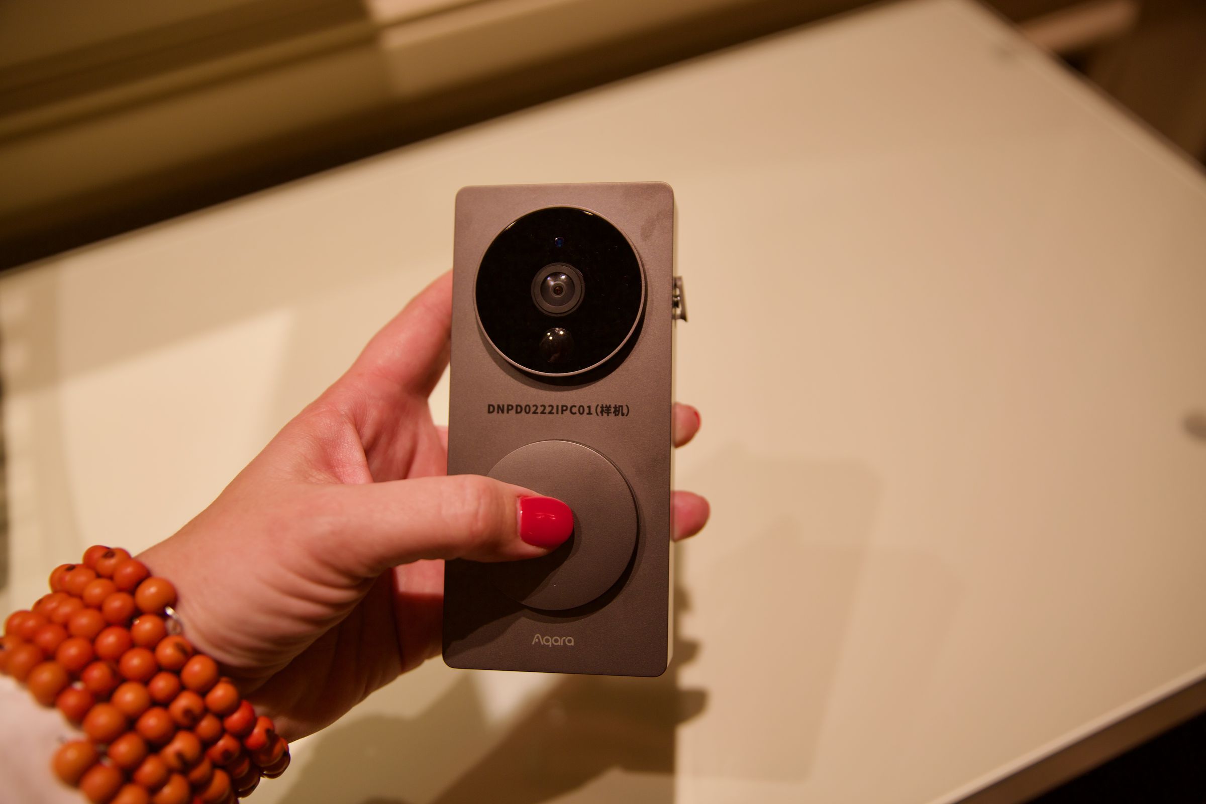 A hand holding a black video doorbell over a table.