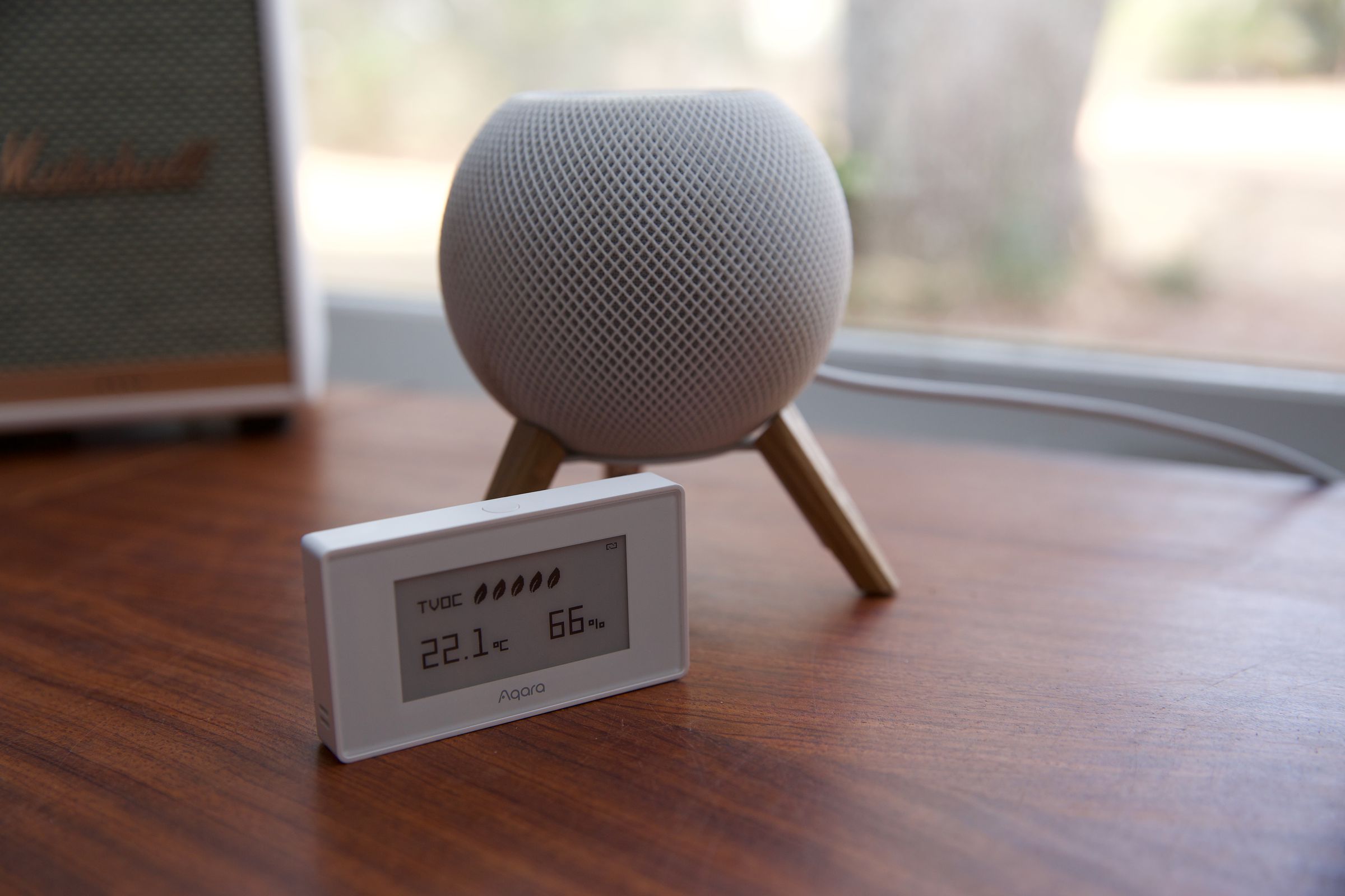 A white HomePod Mini on a desk with a white sensor with an LCD screen next to it.
