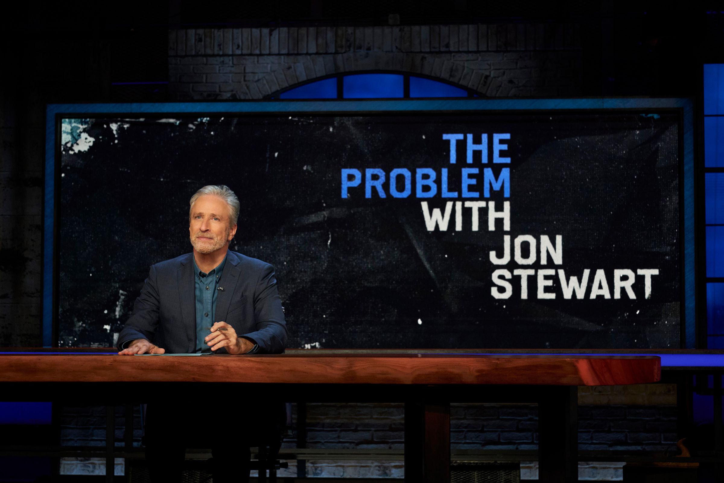 A wide of of a man wearing a suit jacket and shirt sitting at a long table in front of a screen that displaying the words “The Problem With Jon Stewart.)
