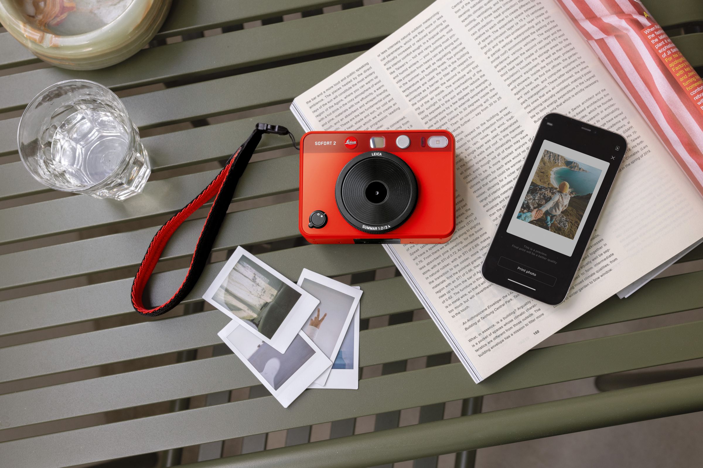 The red Leica Sofort 2 lying on a table against an open book with some photos and a smartphone. 
