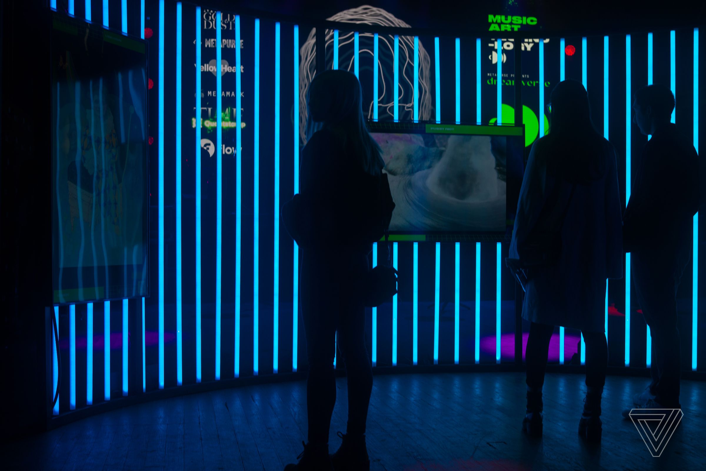 People stand in front of tubes of TVs showing NFT artwork. The TVs are mounted against tubes of blue light.