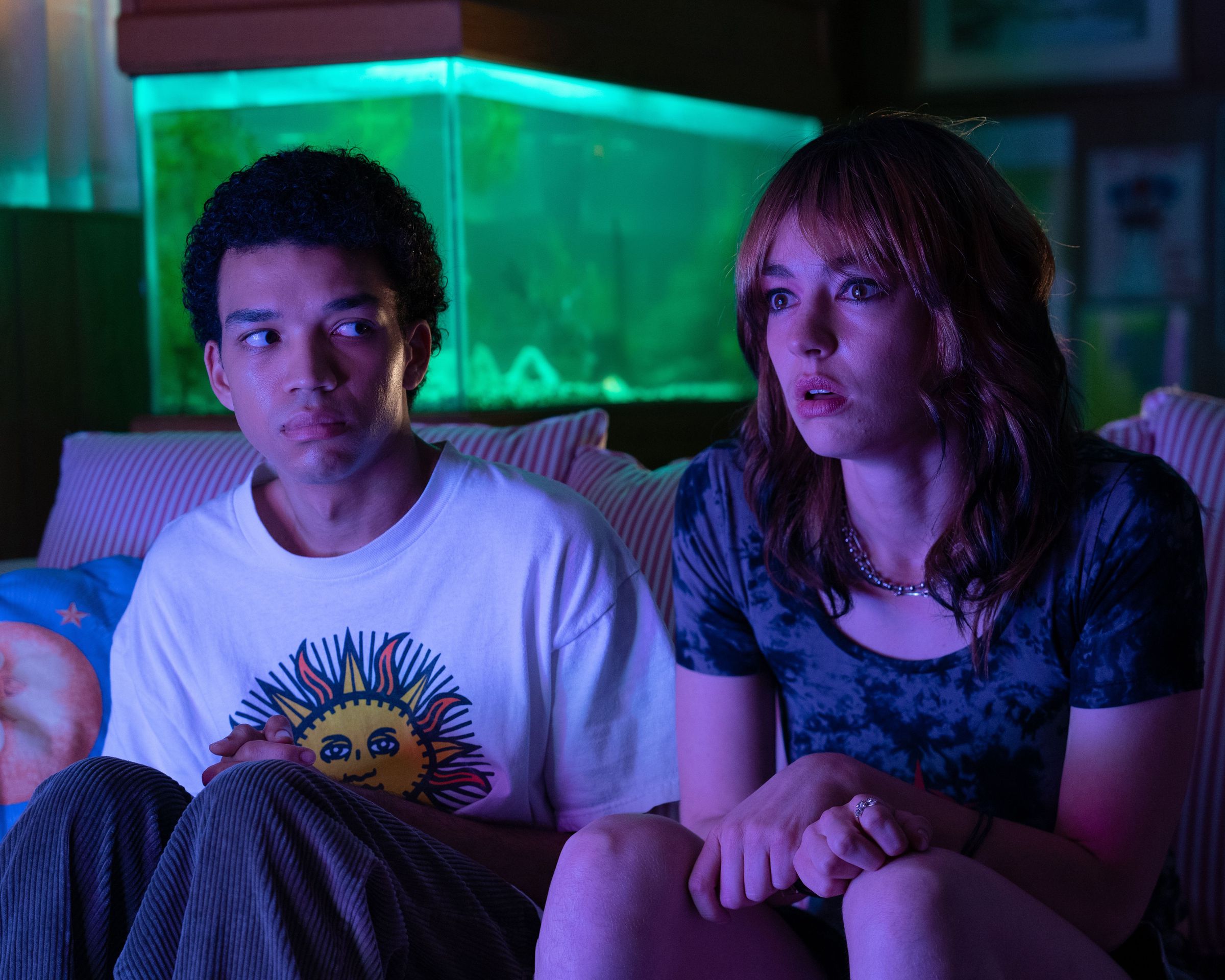 A young man in a t-shirt and pajama bottoms sitting next to a young woman in a t-shirt and shorts. The pair are sitting on a couch in front of a glowing television, behind them stands a fish tank.