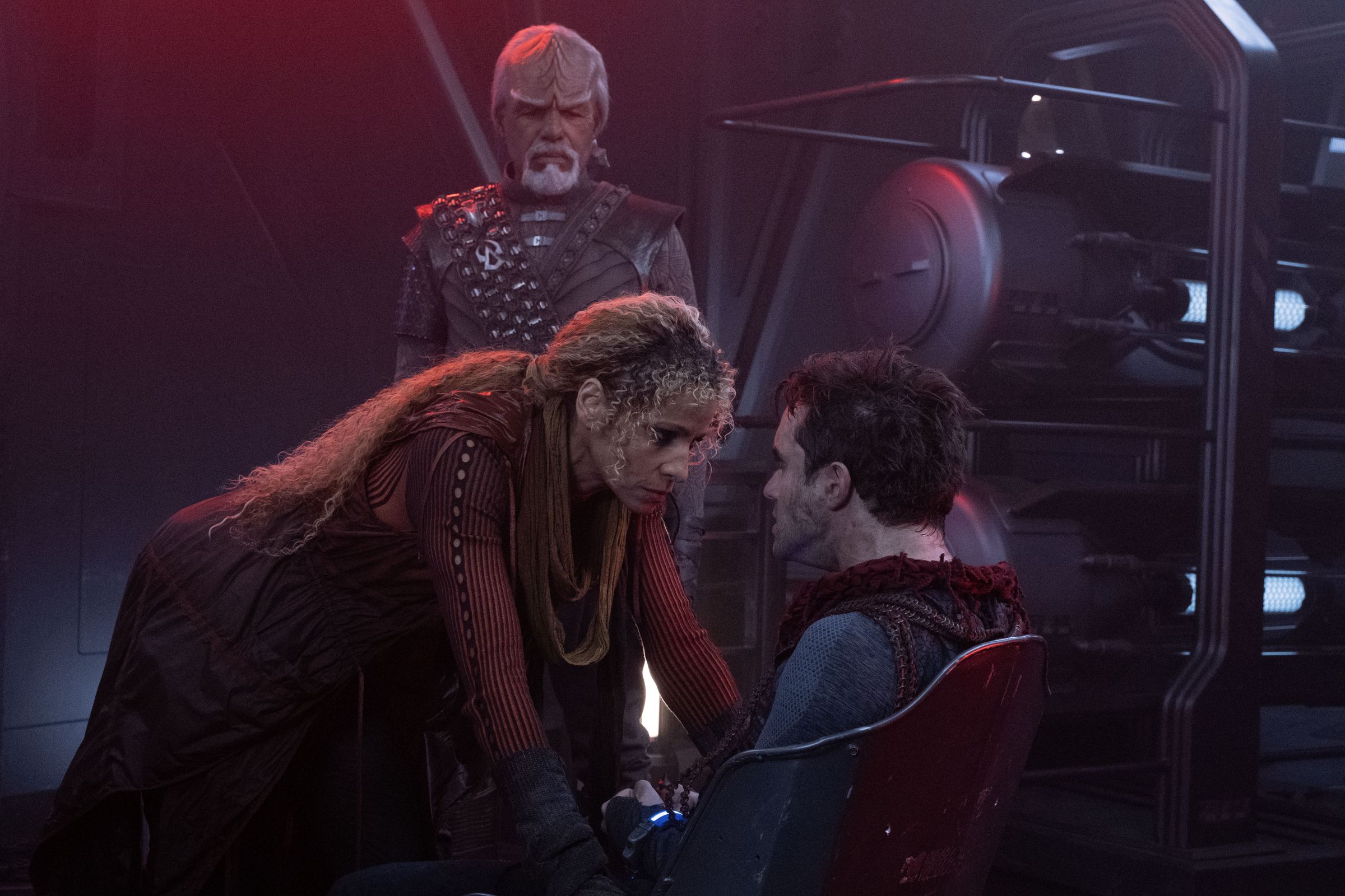 Worf, an older male Klingon, and Raffi, a Black woman with blond hair, interrogate a greasy white male villain.