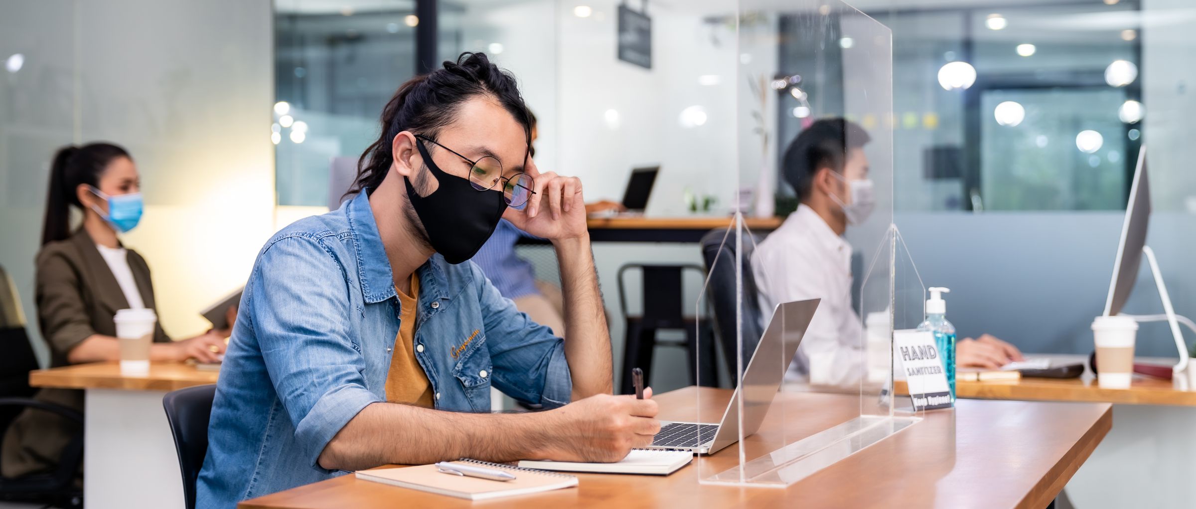 Workers wear protective face masks in a socially distant office, with hand sanitizer at their desks. 