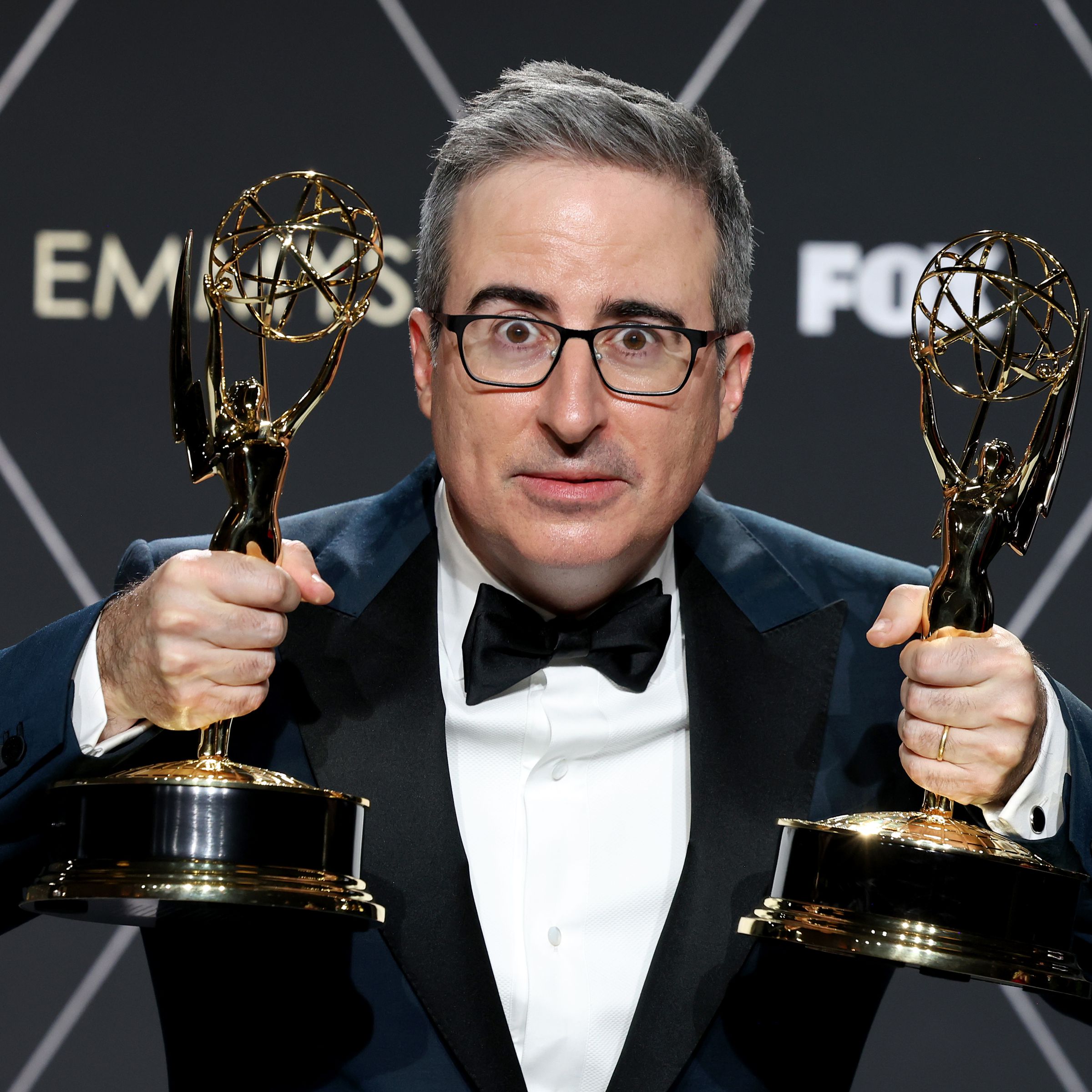 John Oliver wearing a Tuxedo and making a distraught face while holding two Emmys.
