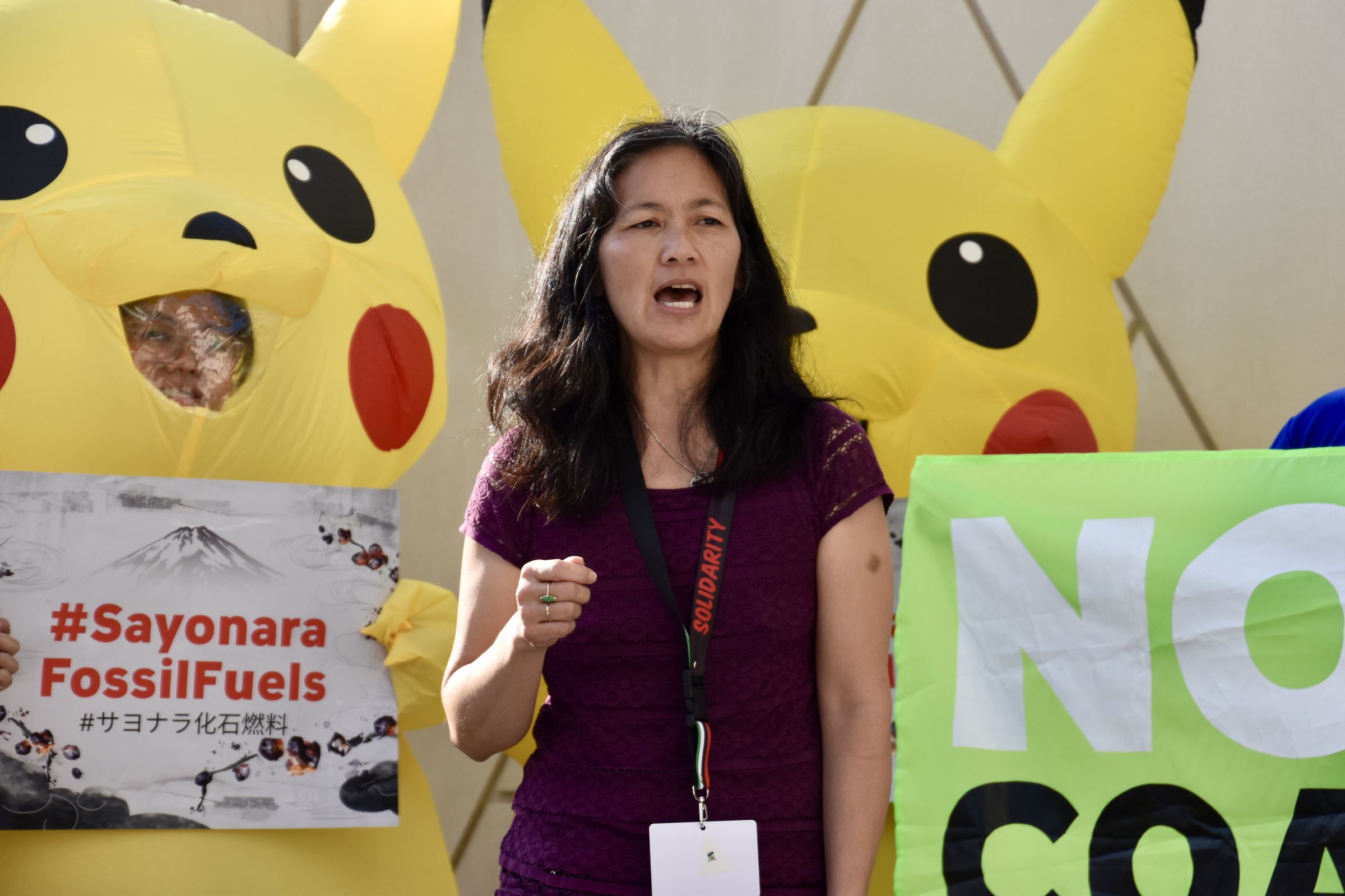Two people wearing inflatable Pikachu costumes stand side by side behind a person speaking. One holds a sign that says “#Sayonara FossilFuels”.