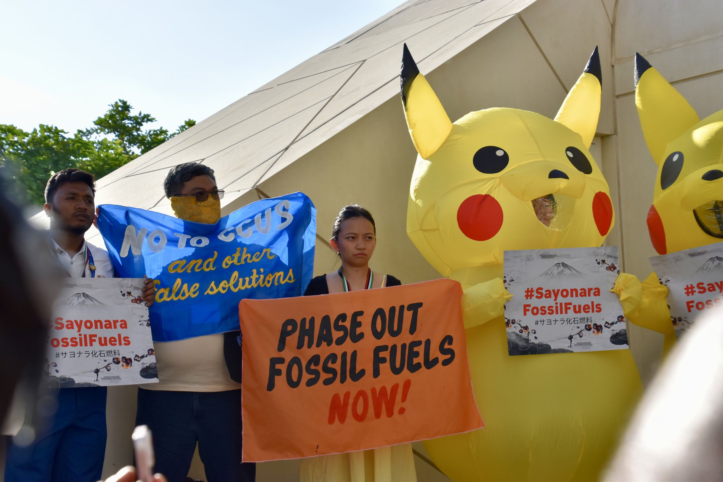 Two people wearing inflatable Pikachu costumes stand side by side holding signs that say“#Sayonara FossilFuels”. They stand next to other demonstrators holding banners that say “phase out fossil fuels now!” and “No to CCUS and other false solutions”.
