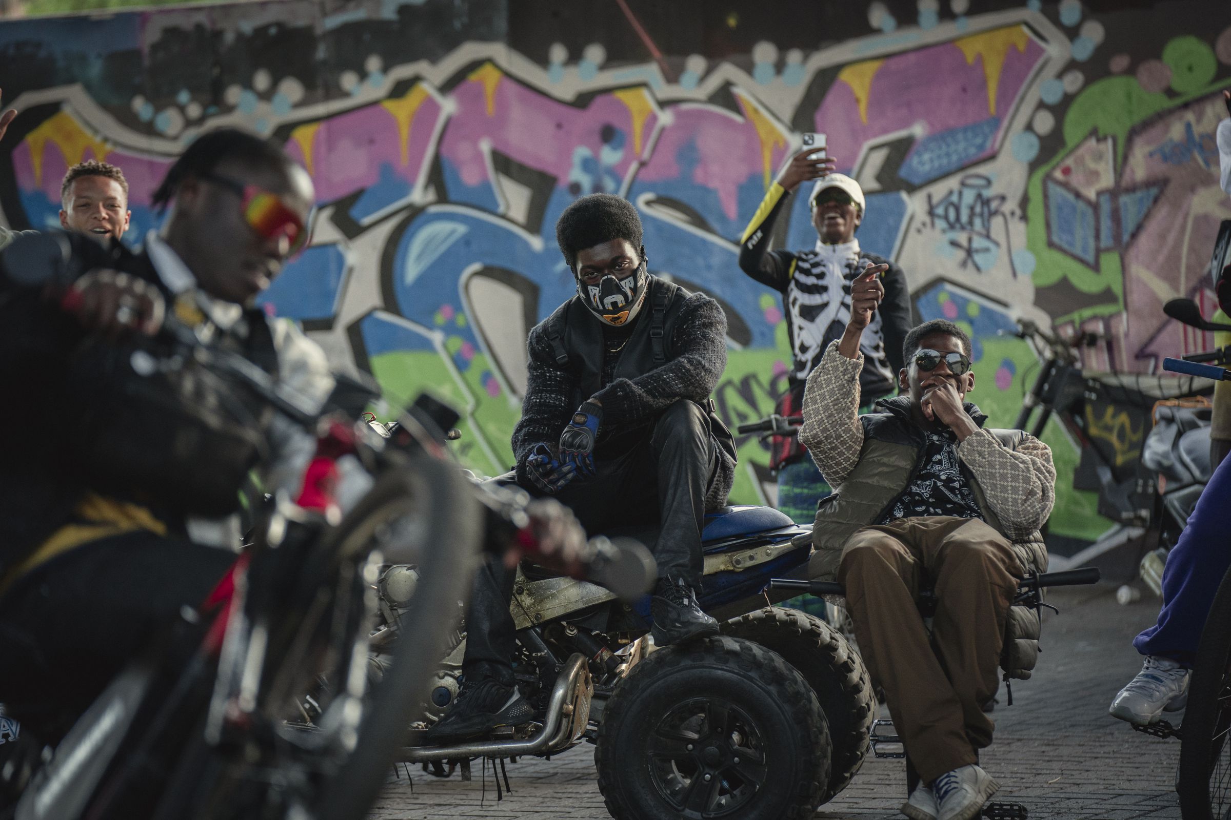 A man wearing a motorcycle mask, black jackets, pants, and boots sitting on top of an ATV. Around the man is an assortment of other people sitting on motorized bikes and motorcycles doing tricks, and behind the group is a massive wall covered in street art.