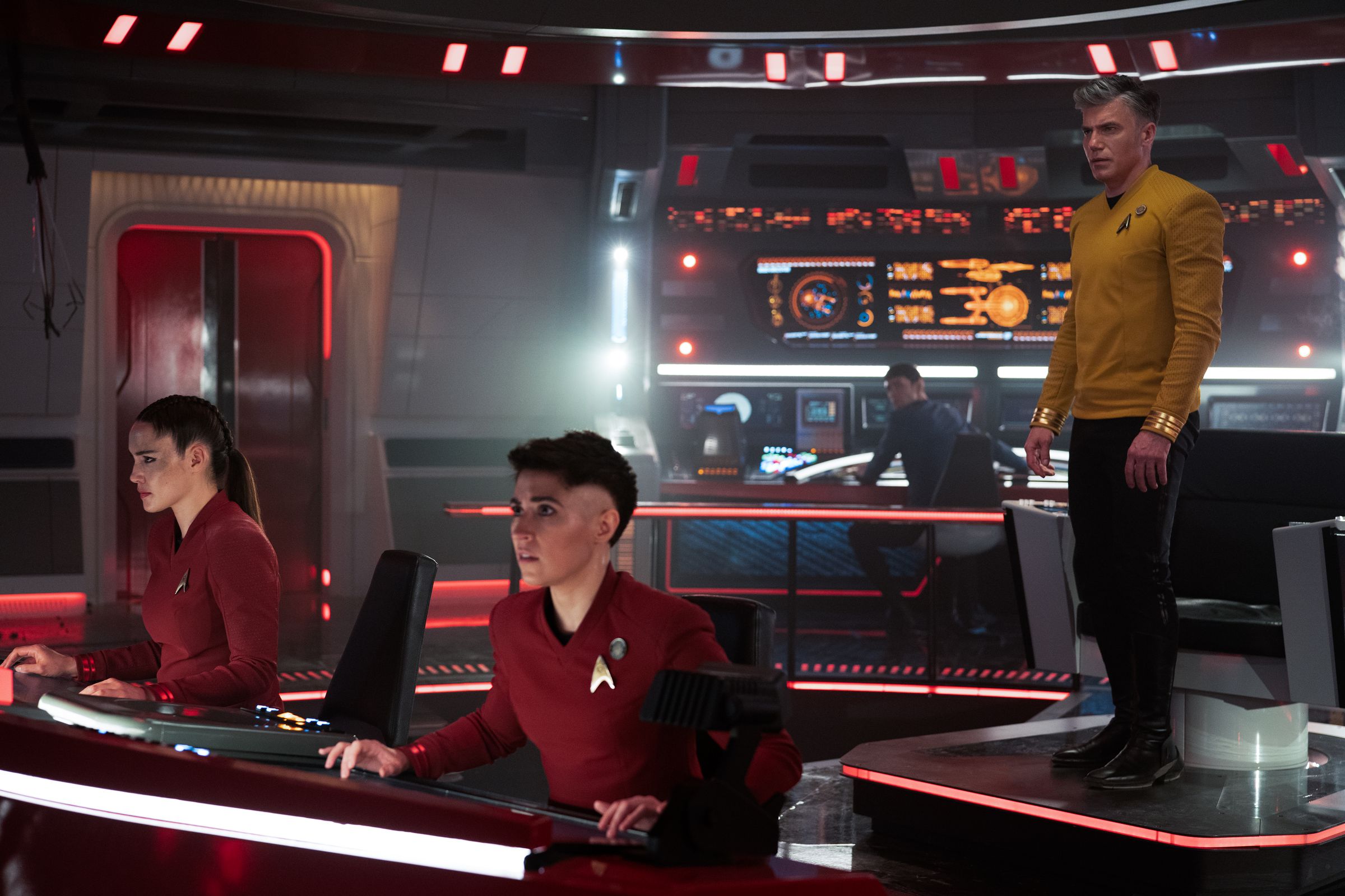 A gray-haired White man wearing yellow stands and looks at something off screen. Two women with dark hair and red shirts are seated and looking in the same direction.