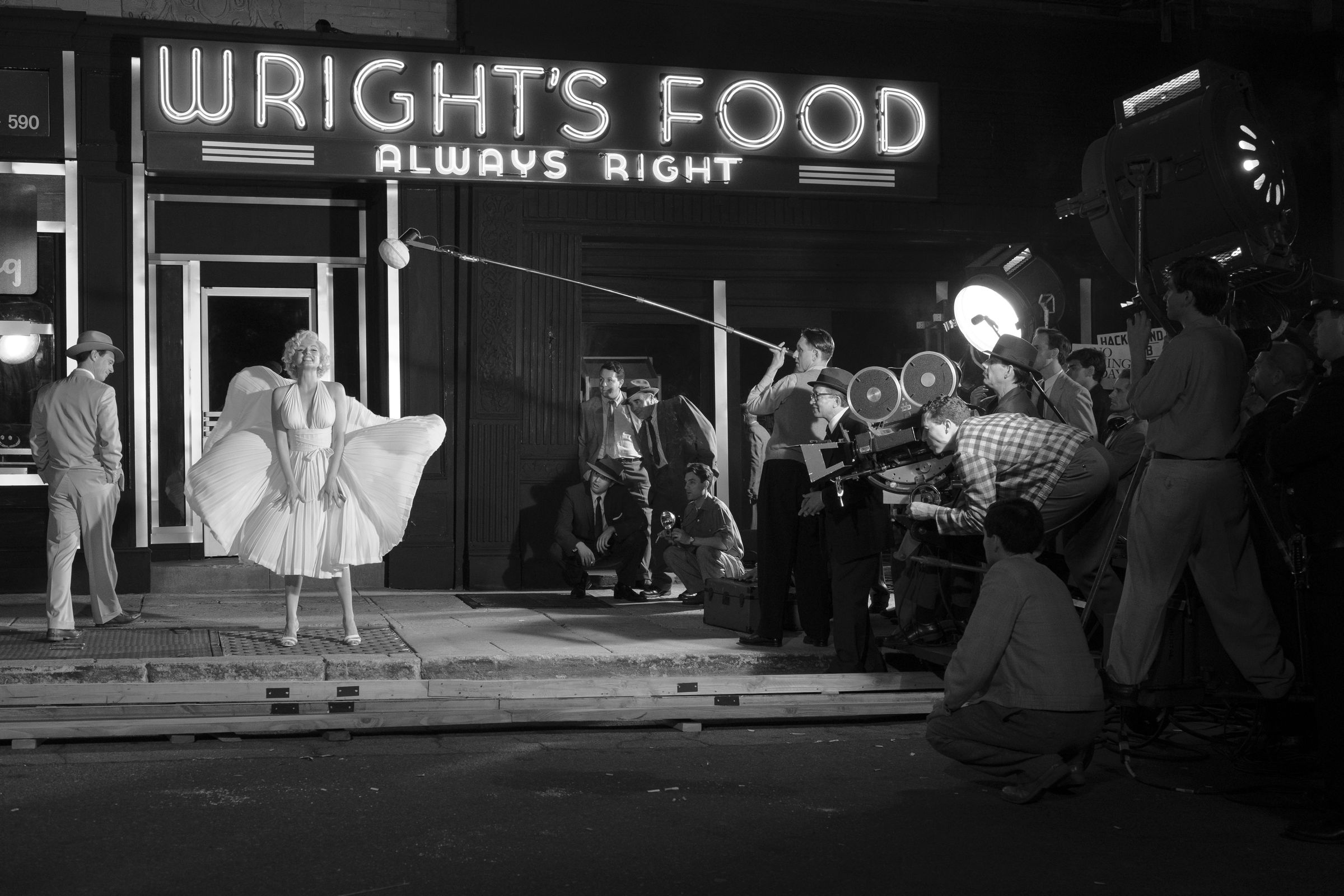 A black and white recreation of the iconic scene from The Seven Year itch in which a woman in a white cocktail dress stands on a grate on a sidewalk as a train passes by underneath, causing her dress to billow up around her. To the woman’s right is her co-star, a man in a suit looking at her, and to her left stands a the massive gaggle of men who make up the crew filming the scene.