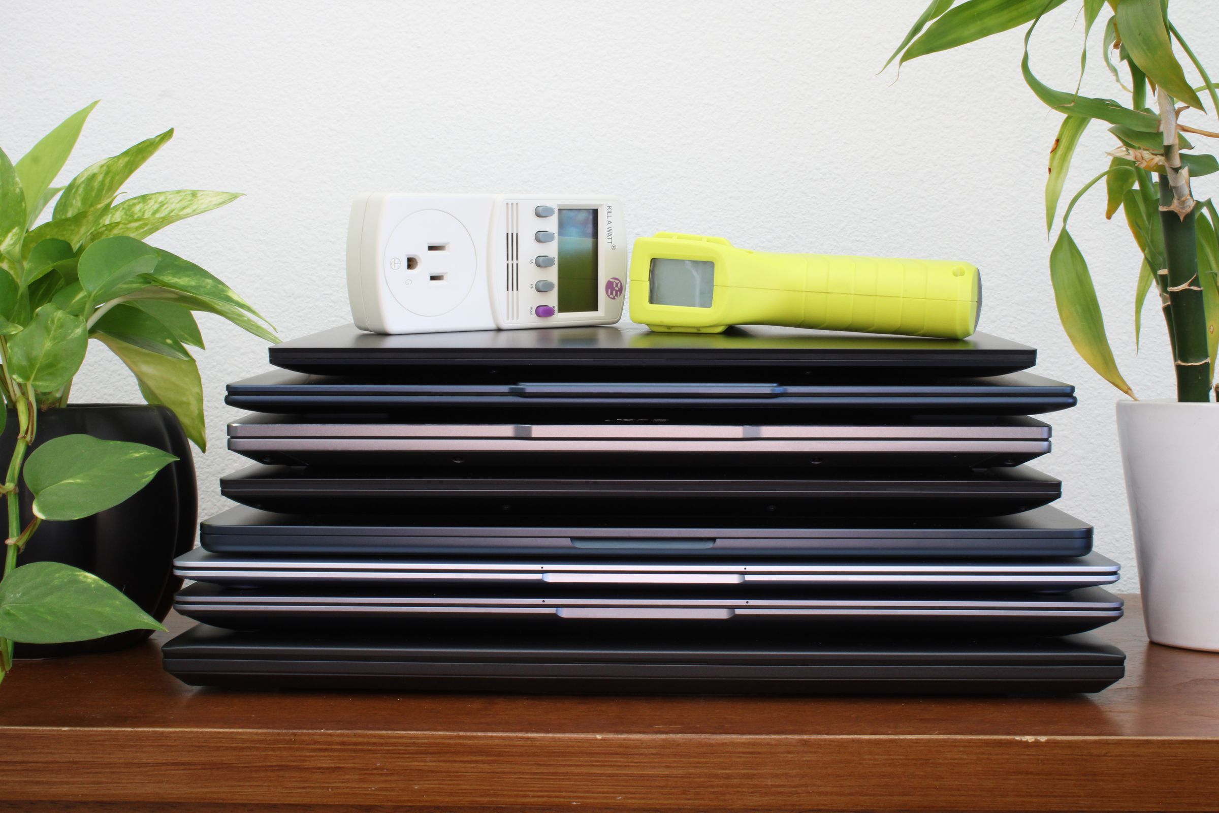 A stack of laptops on top of a brown table, flanked by two green plants, against a white wall.