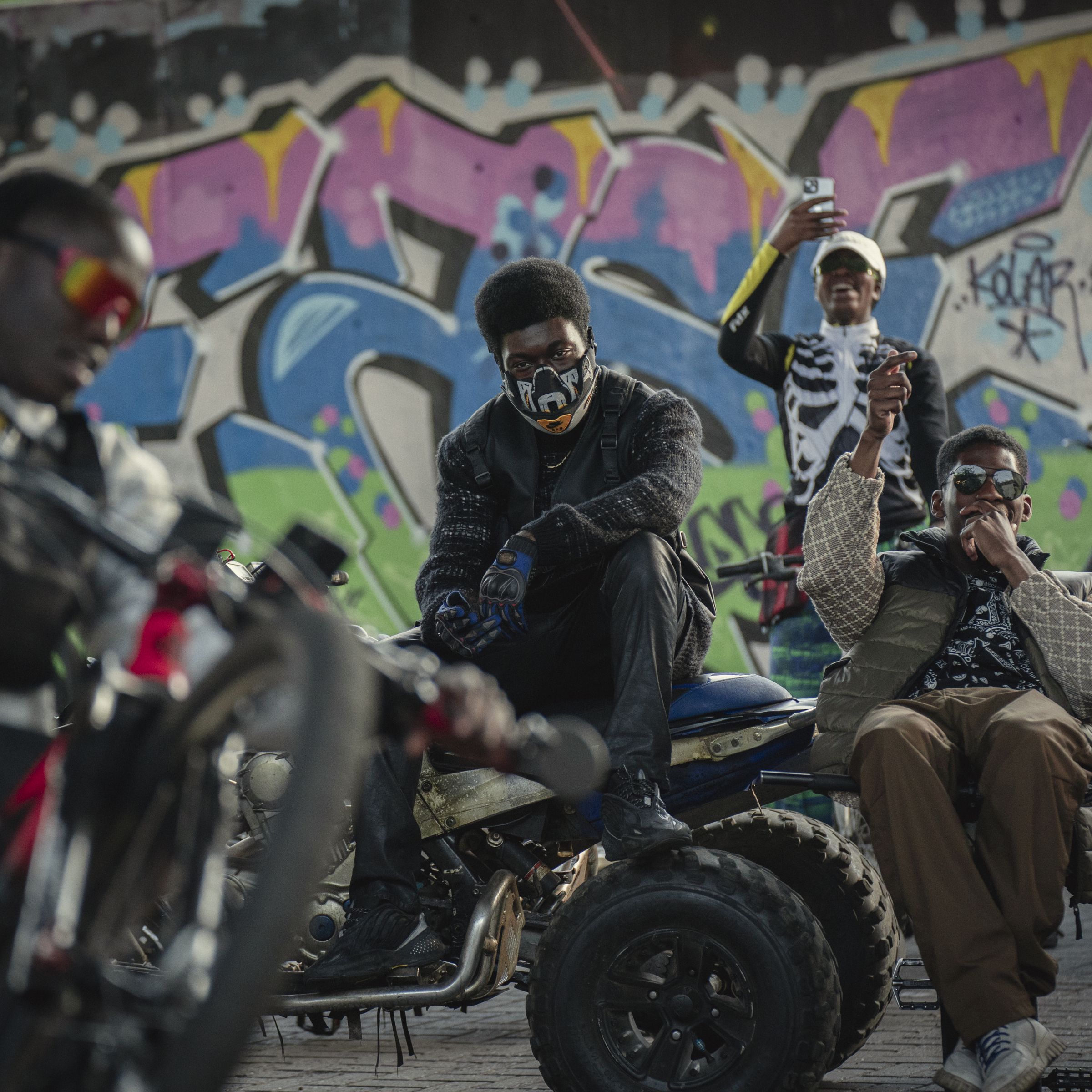 A man wearing a motorcycle mask, black jackets, pants, and boots sitting on top of an ATV. Around the man is an assortment of other people sitting on motorized bikes and motorcycles doing tricks, and behind the group is a massive wall covered in street art.