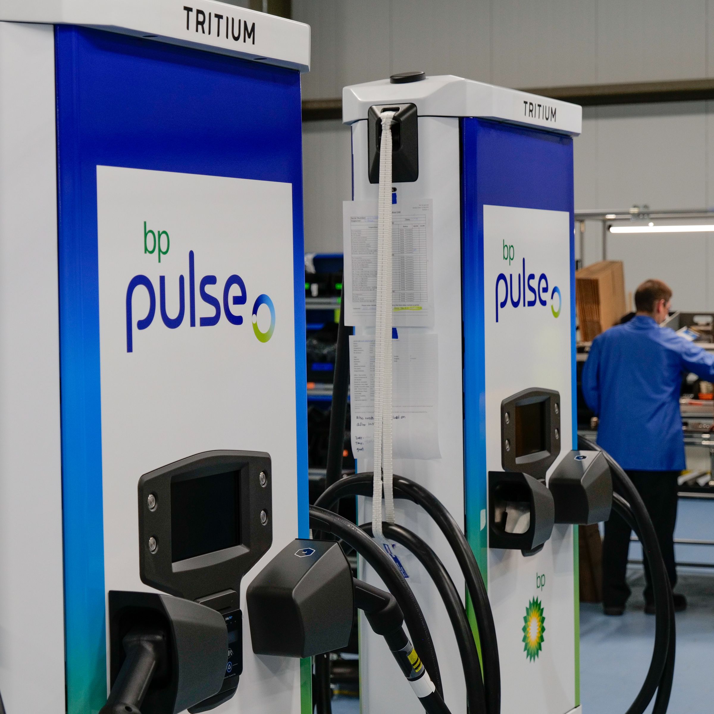 Two charging stations are in a production facility with people working in the background. The chargers have Tritium logos on the top and BP Pulse logos on the center, and each have two charging plugs.