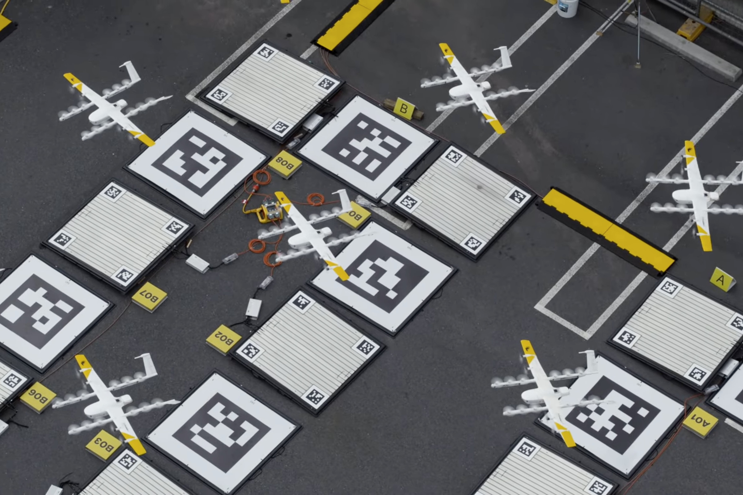 rooftop with squares, some with QR codes, others for drones to land to charge, 6 drones are lifting off from the pads.
