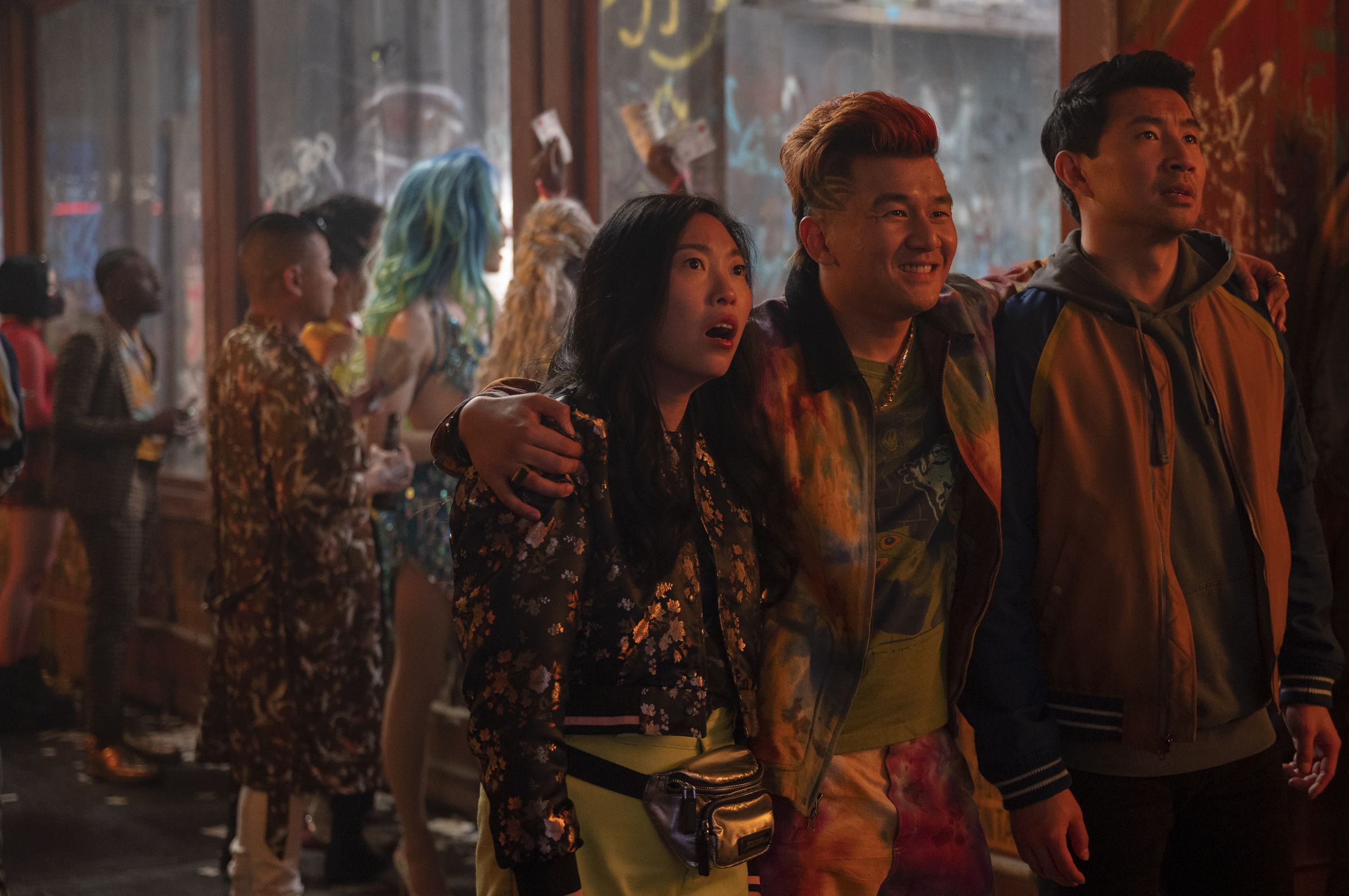 Awkwafina as Katy, and Simu Liu as Shang, stare in open mouthed shock at something out of frame as Ronny Chieng as Jon Jon embraces them both while looking amused.