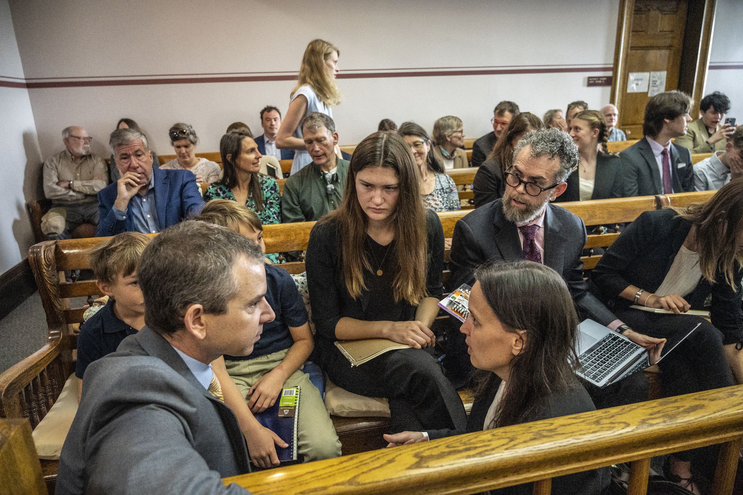 A young woman sits in a crowded courtroom with a team of lawyers around her.