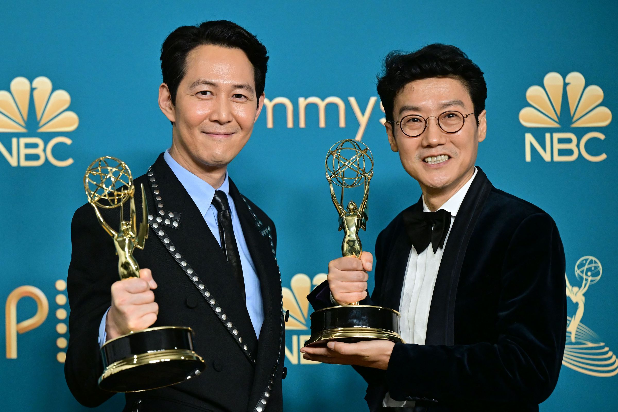 Actor Lee Jung-jae and Squid Game director Hwang Dong-hyuk posing with their Emmys in front of a turquoise backdrop.