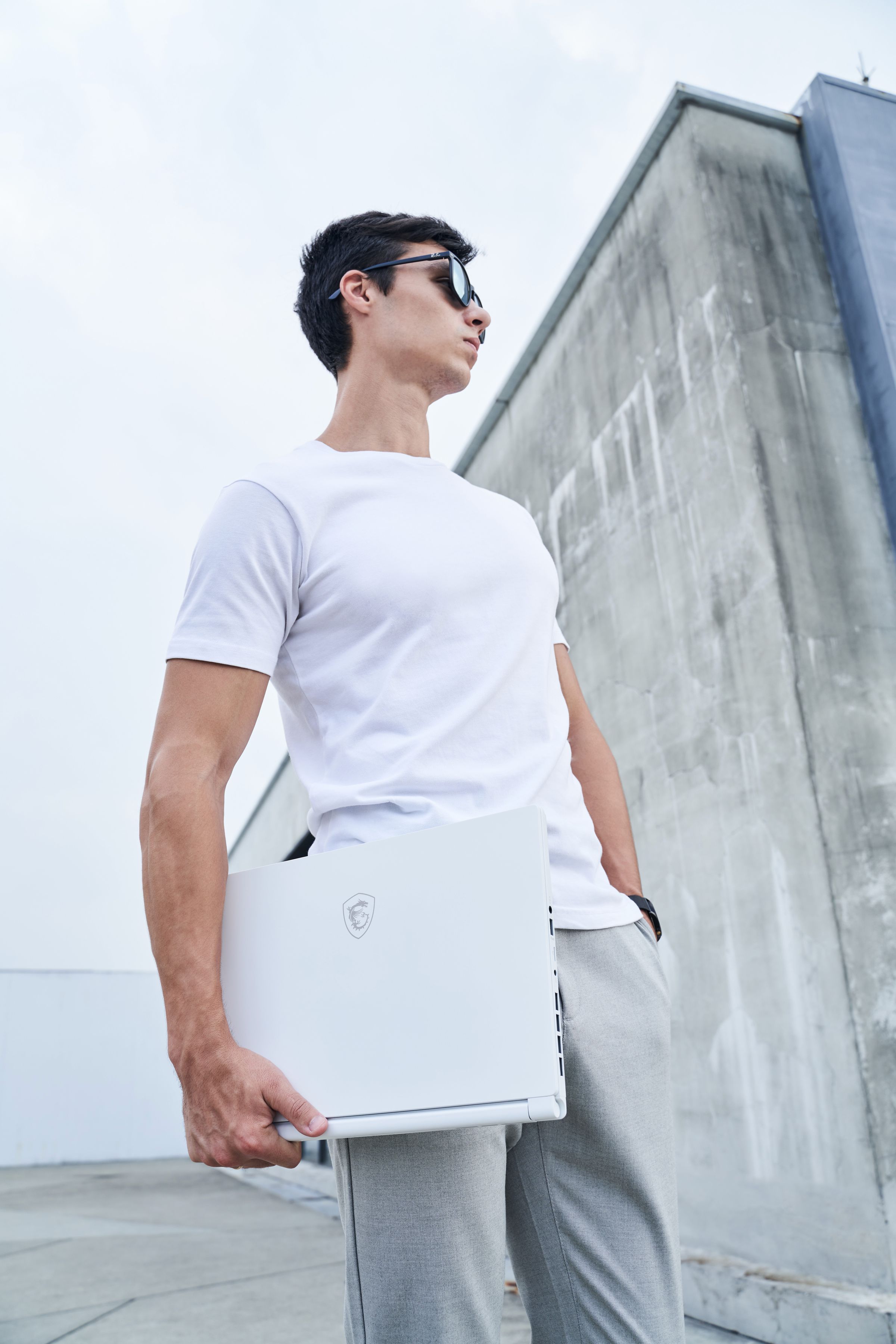 A user stands on the roof of a building wearing a white T-Shirt, sunglasses and sweatpants. They carry a white MSI Stealth 15M under their right arm while looking off to the left.