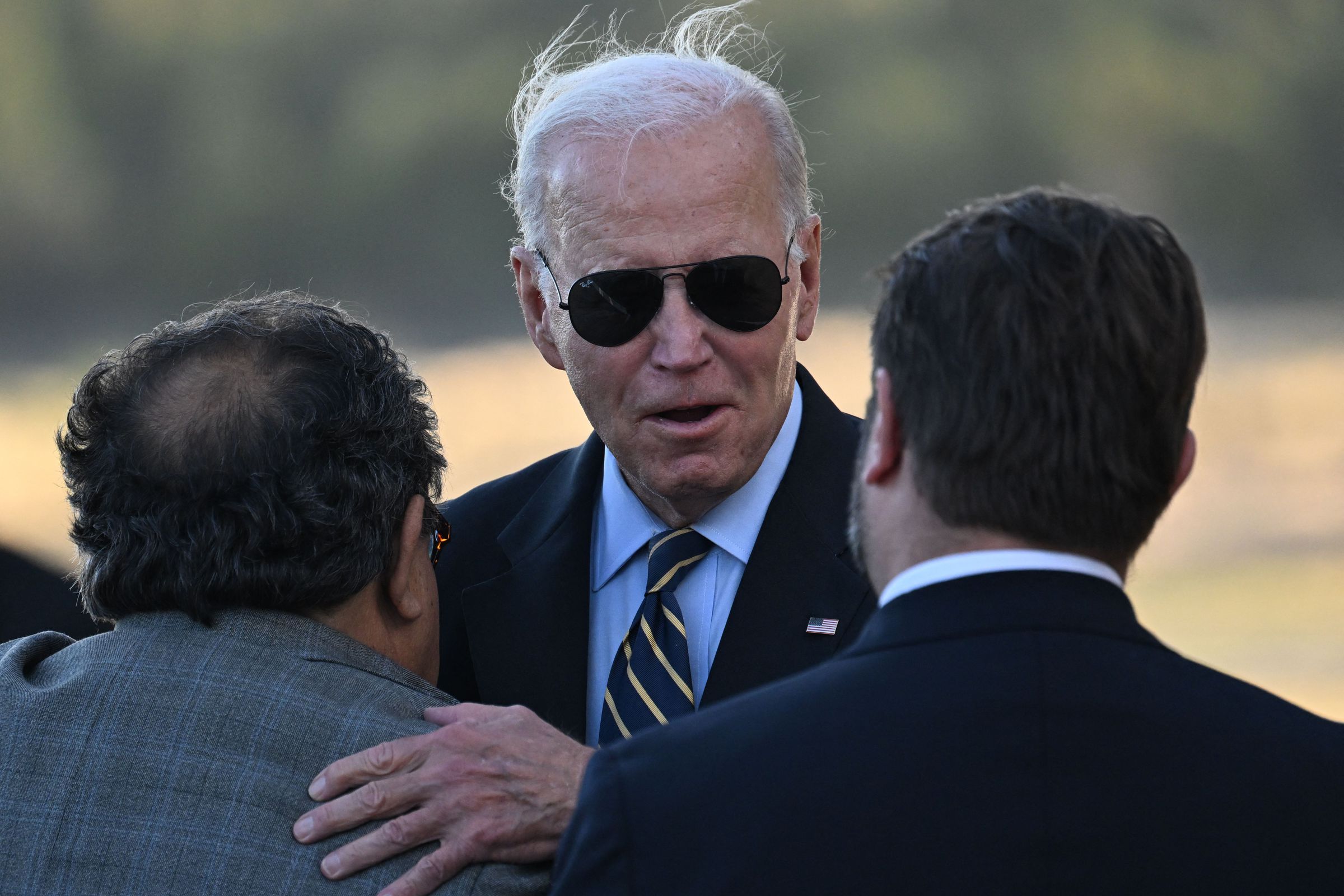 Joe Biden wearing sunglasses and greeting two other people in suits.
