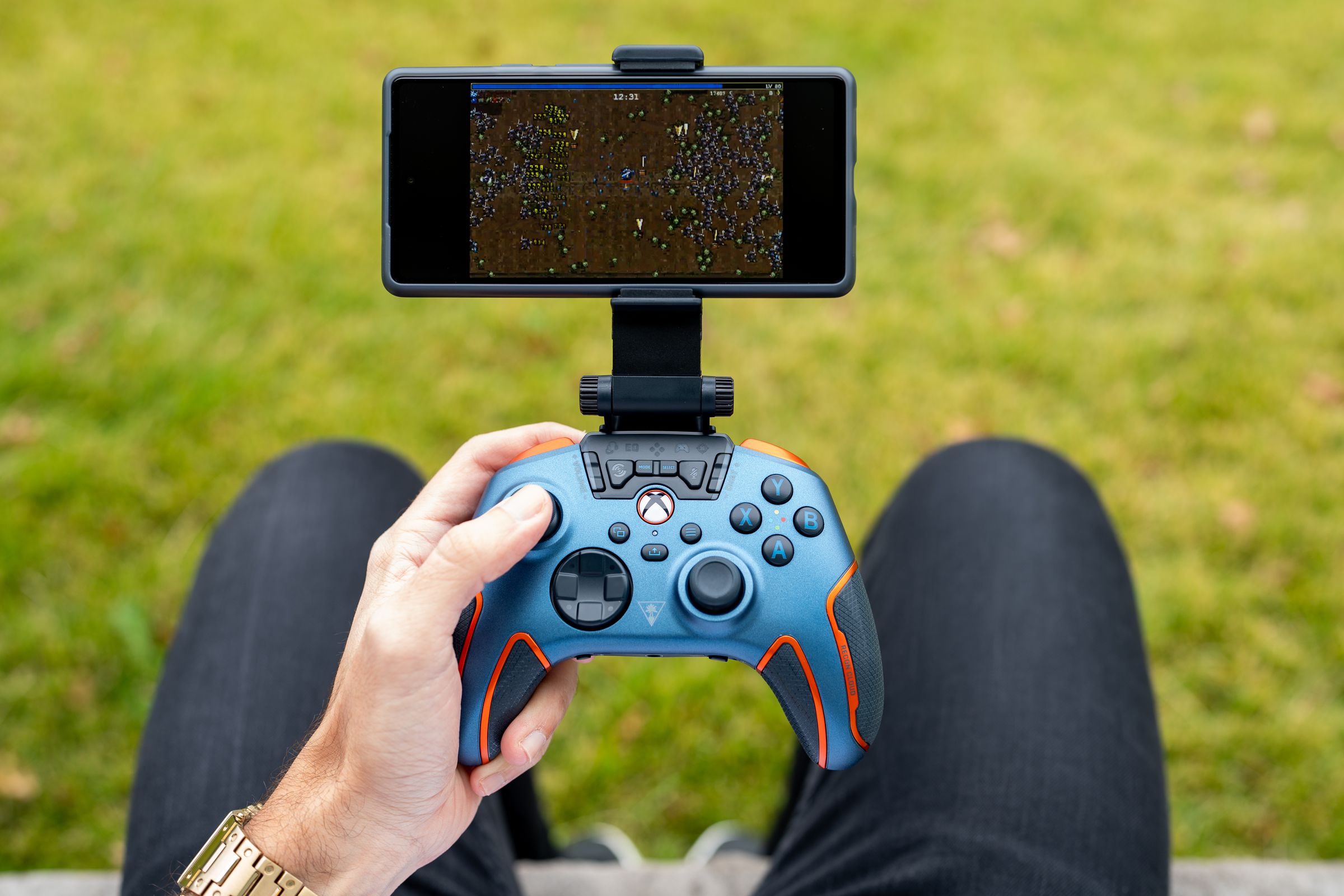 A person holding the blue and orange Turtle Beach Recon Cloud controller above their lap while outside in front of green grass. The controller has a smartphone mounted to its top, streaming a game from the cloud.