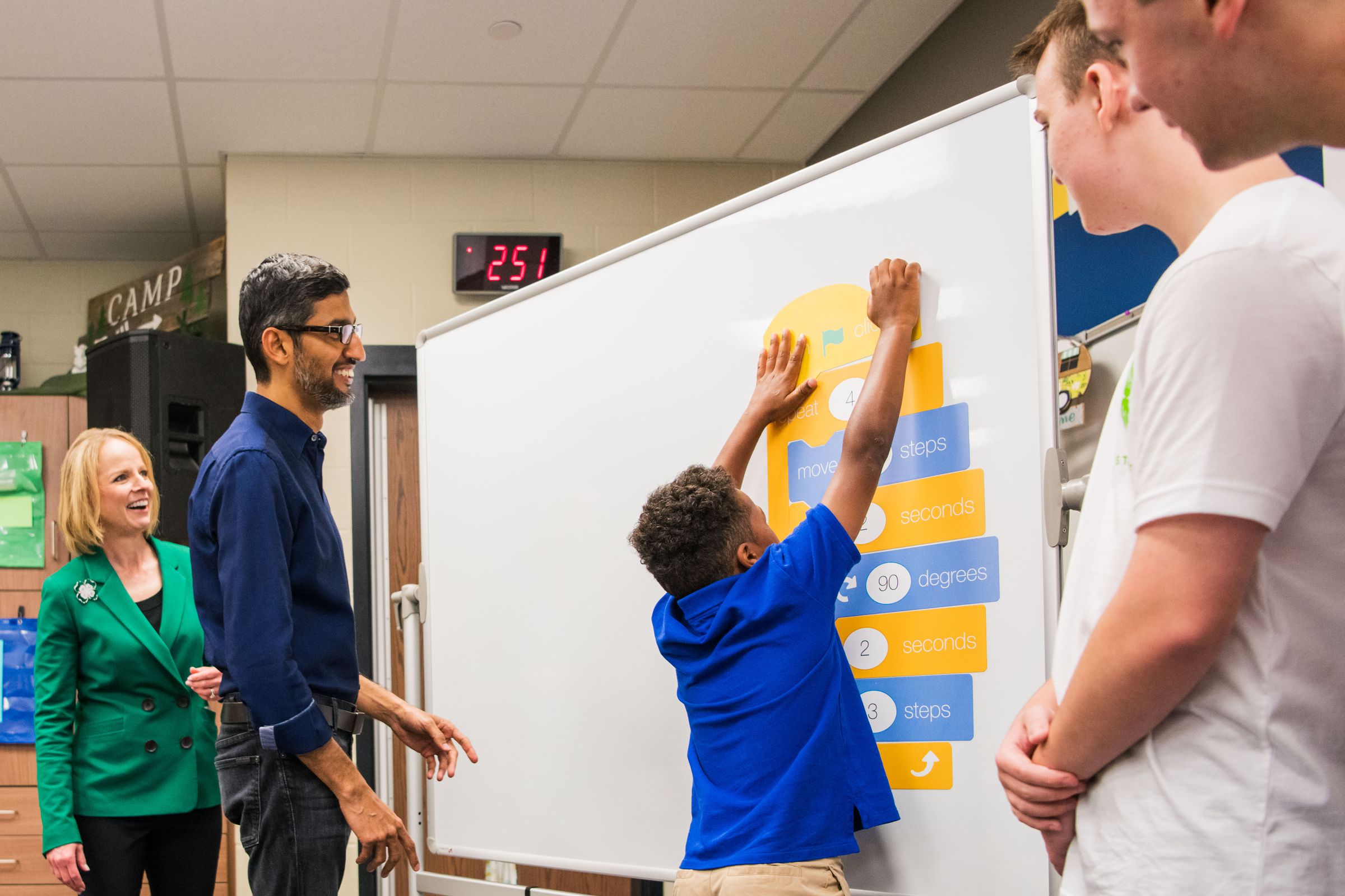 In a classroom, Sundar Pichai and Jennifer Sirangelo watch students place stickers on a whiteboard.