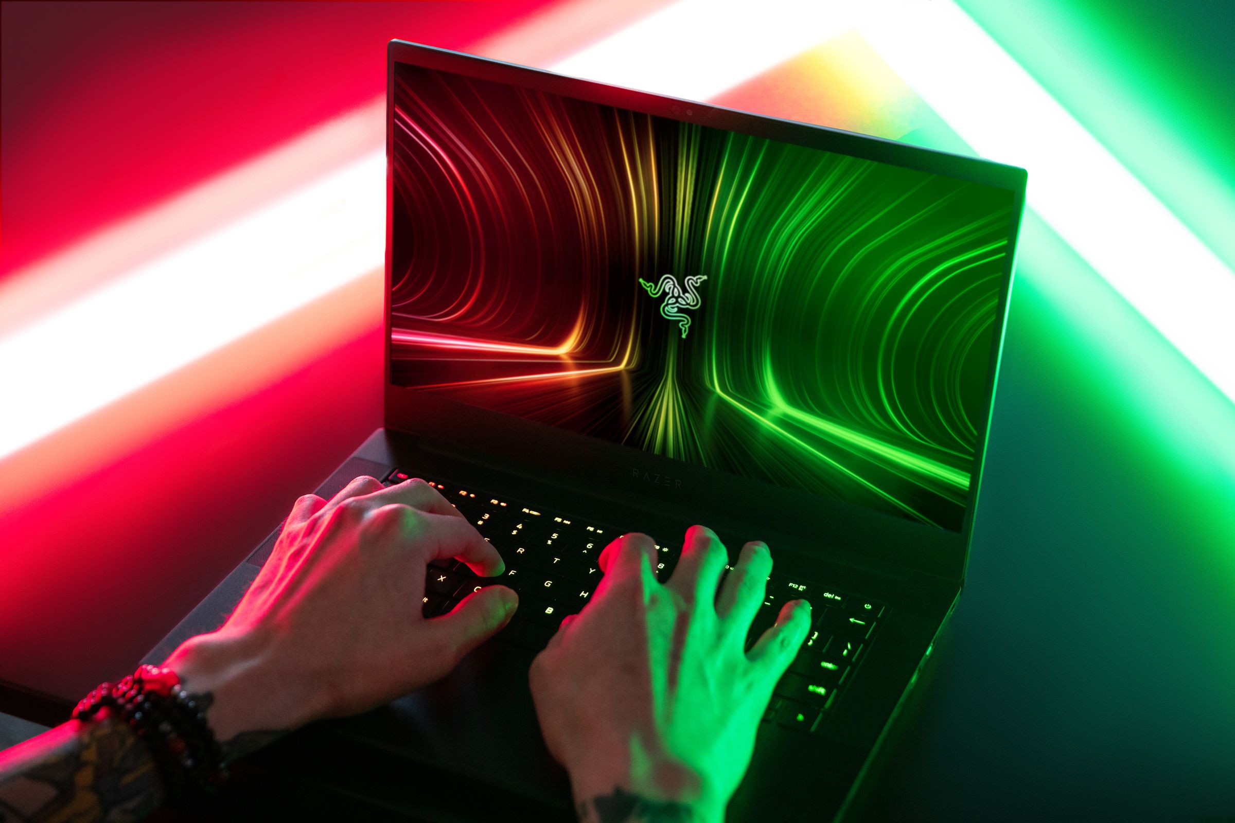 An over-the-shoulder shot of a user typing on the Razer Blade 14. The screen displays the Razer logo on a red, green, and black background.