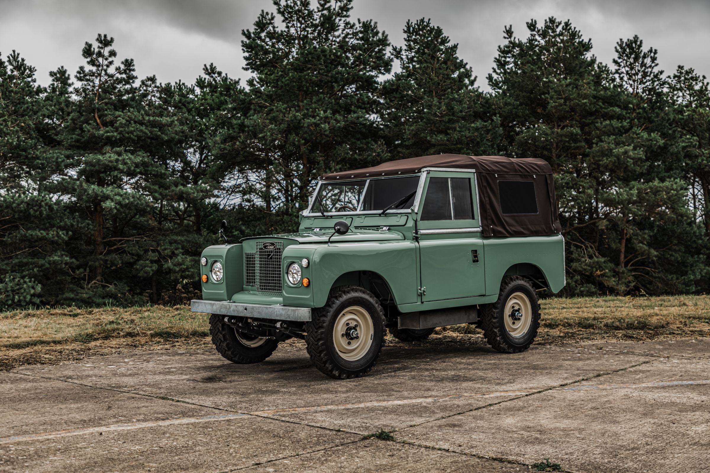 Green boxy Land Rover vehicle with a cloth top on a battered concrete slab surrounded by trees