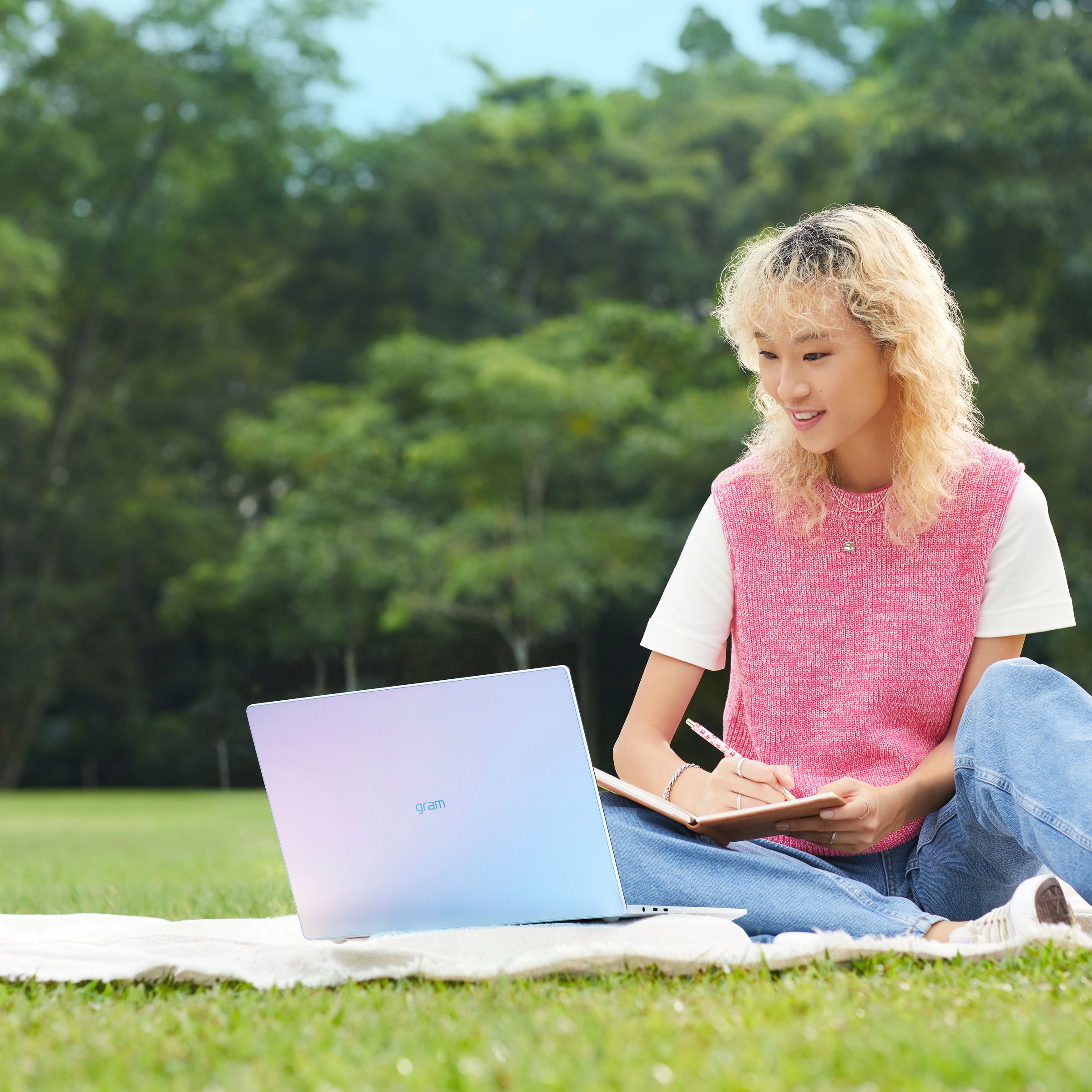 A user writes in a notebook while sitting on a picnic blanket in a park with an LG Gram Style model and a drink.